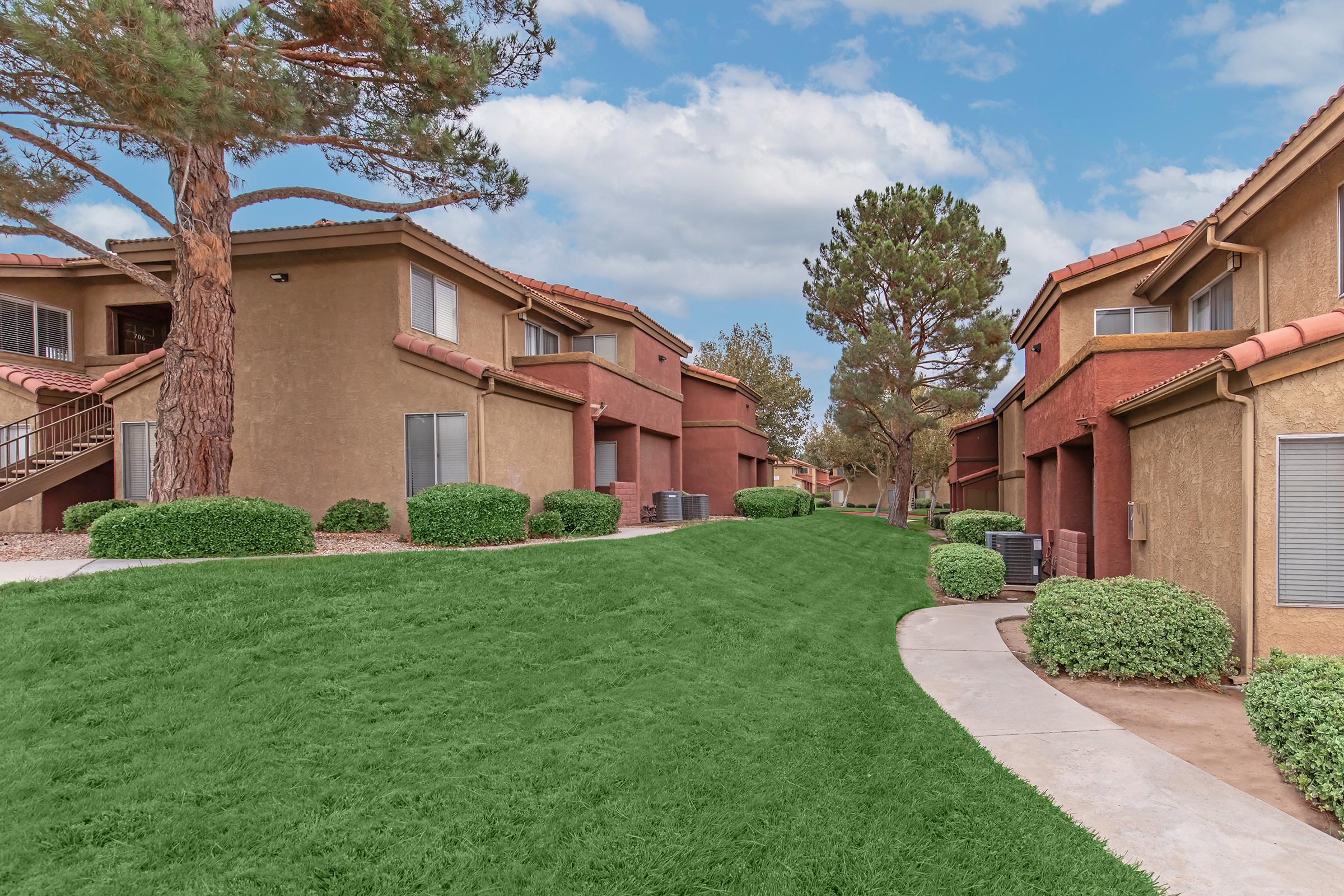 A landscaped pathway winds through a residential area, featuring two-story buildings with brown exteriors. Lush green grass and neatly trimmed bushes line the pathway, while trees provide shade under a partly cloudy sky. The scene conveys a peaceful and well-maintained living environment.