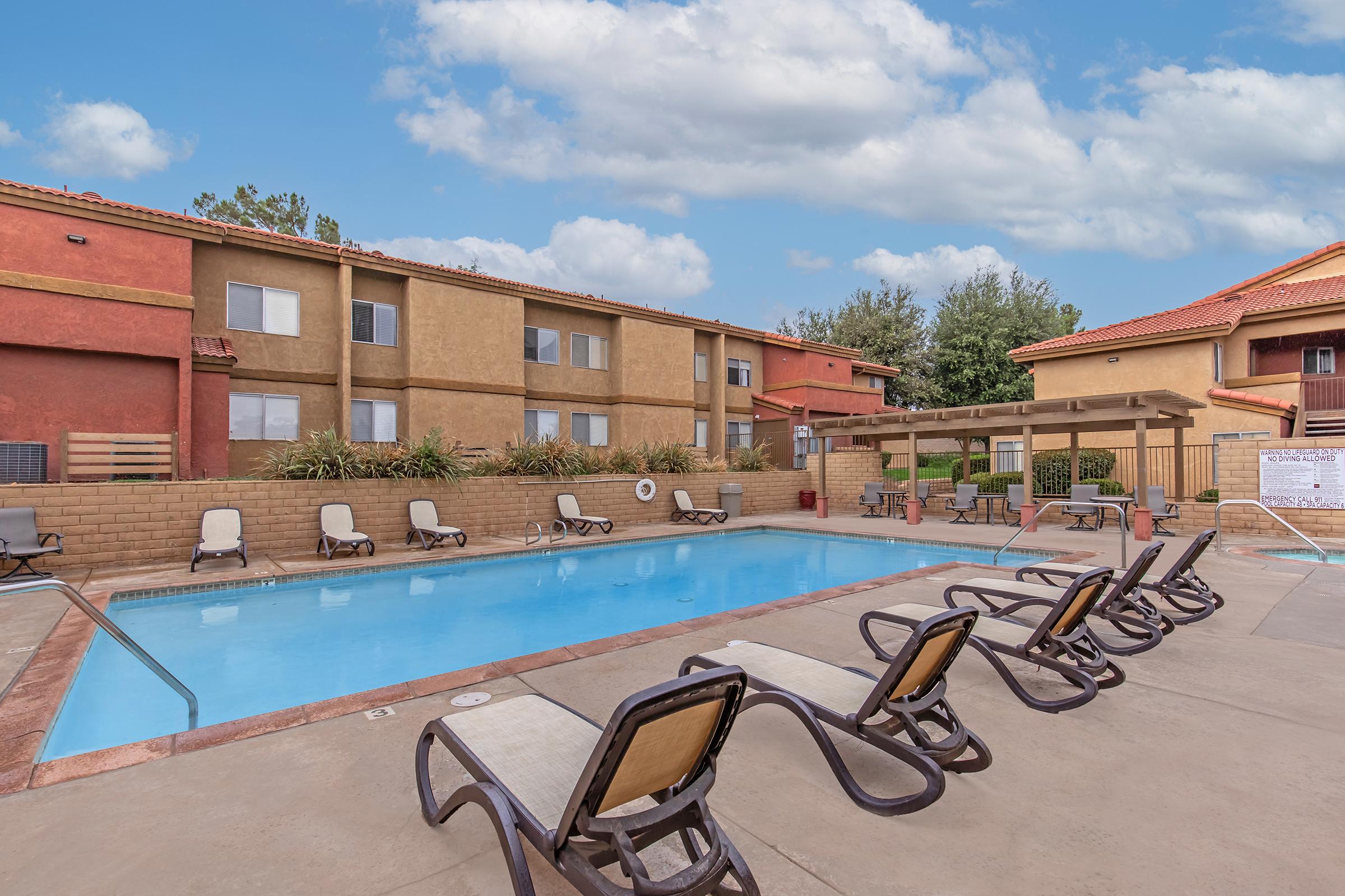A clear blue swimming pool surrounded by lounge chairs, with a shaded seating area nearby. In the background, there are colorful apartment buildings and lush greenery under a partly cloudy sky.