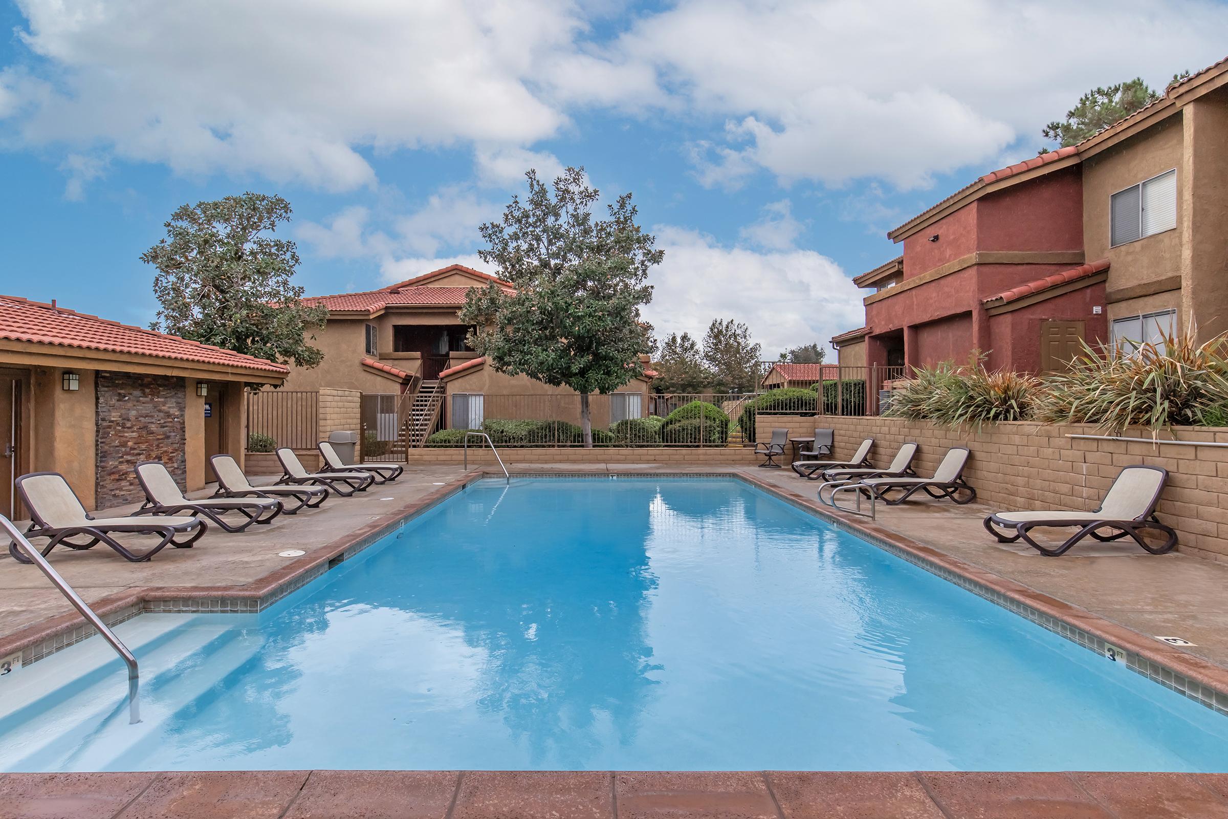 A clear swimming pool surrounded by lounge chairs, with a backdrop of two multi-story apartment buildings and green trees. The sky is partly cloudy, creating a serene and inviting atmosphere.