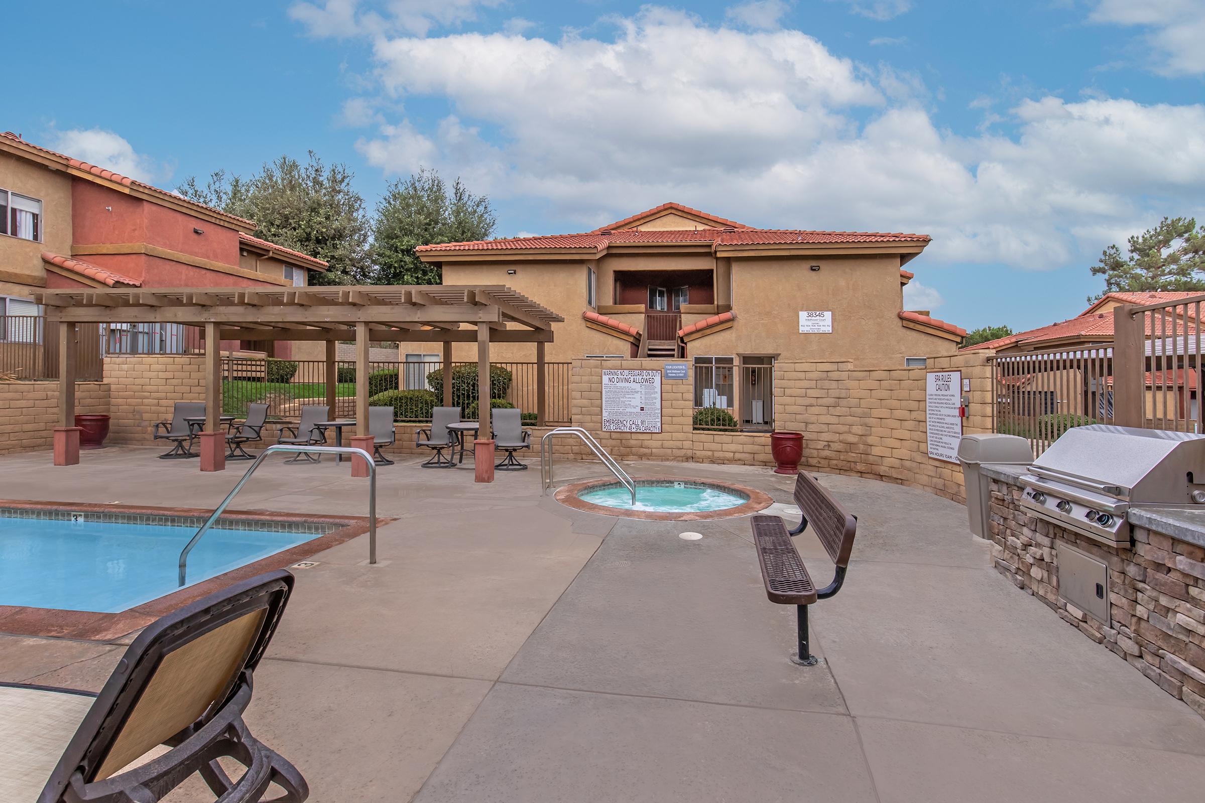 A swimming pool area featuring a jacuzzi, lounge chairs, and a shaded seating structure. The backdrop includes an apartment building with a tan exterior and red tile roof. The scene is brightened by a partly cloudy sky, creating a relaxing atmosphere for residents.