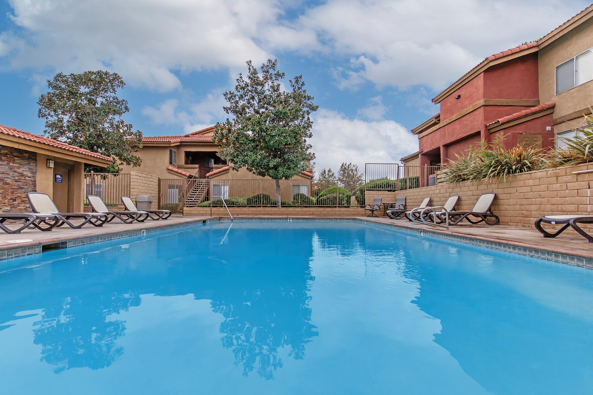 A serene swimming pool surrounded by lounge chairs, with well-maintained landscaping and neighboring buildings featuring red and beige exteriors. The sky is partly cloudy, adding to the inviting atmosphere of a relaxing outdoor space.
