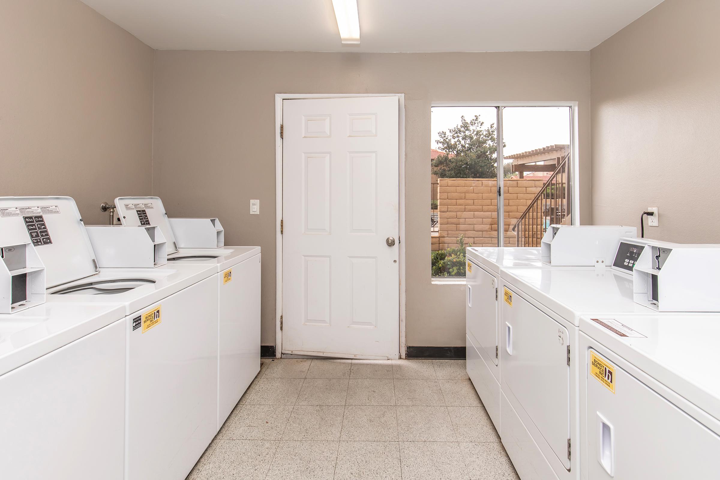A clean, well-lit laundry room featuring several white washing machines and dryers lined up against the walls. A door leads to the outside, and a window provides natural light. The floor is tiled, and the walls are painted a neutral color, creating a tidy and functional space for doing laundry.