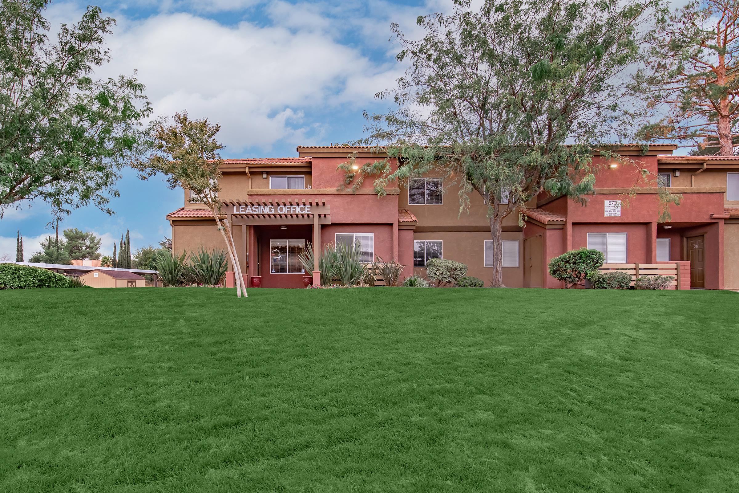 A view of a residential building with a two-story design, featuring tan and brown walls. The building is surrounded by green grass and small shrubs, with trees on both sides. The sky is partly cloudy, creating a pleasant outdoor atmosphere.
