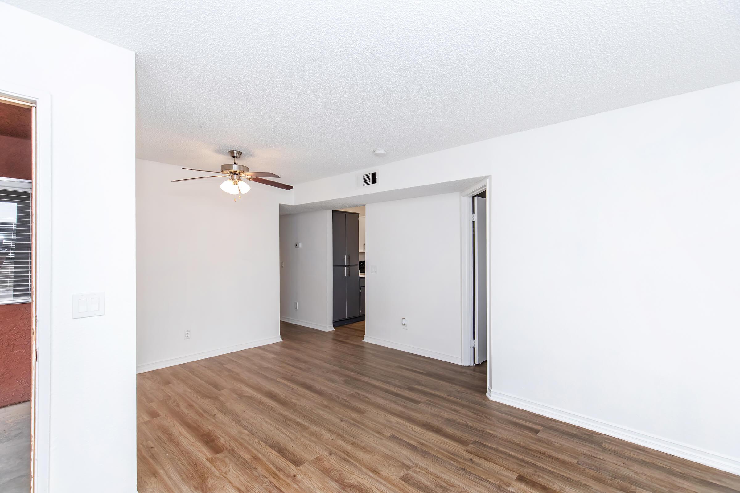 A spacious, well-lit living area featuring light wood flooring, a ceiling fan, and white walls. There are doorways leading to adjacent rooms, with a glimpse of a kitchen area in the background. The overall ambiance is modern and welcoming.