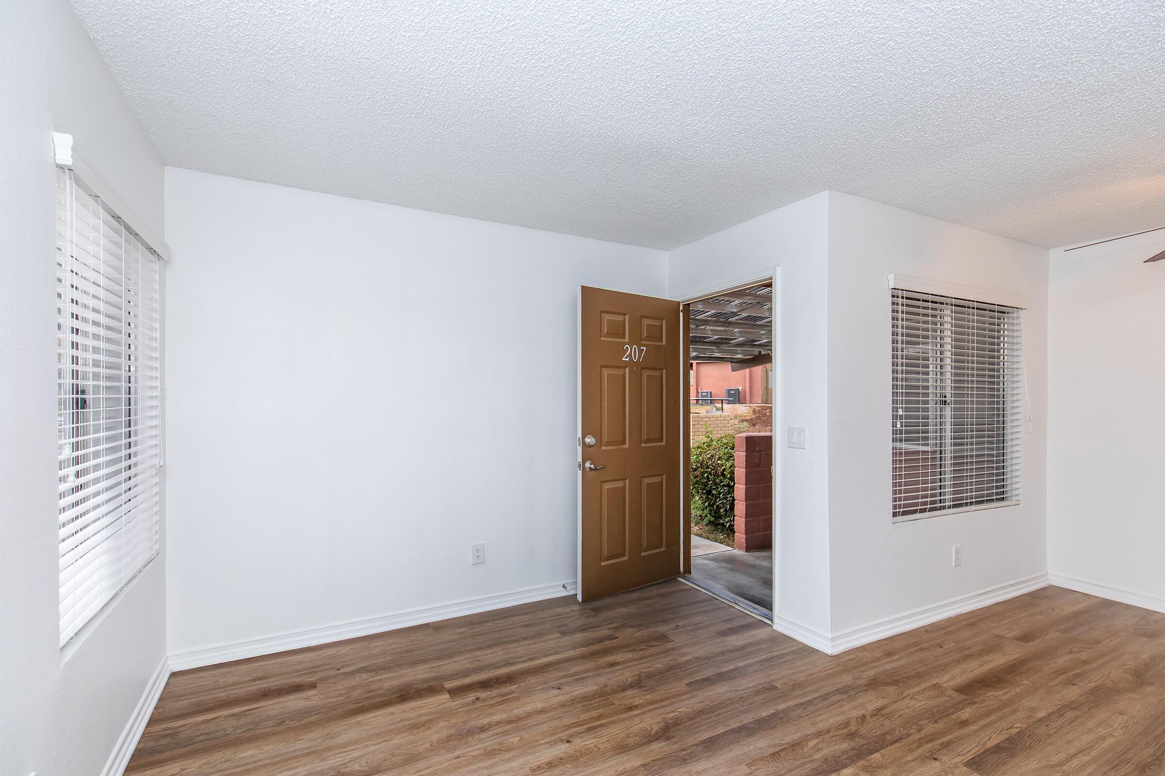 Empty interior view of an apartment room with light-colored walls and wooden flooring. A door, labeled "207," leads to an exterior area, while windows with blinds allow natural light. The space is simple and uncluttered, indicating it is ready for new tenants.
