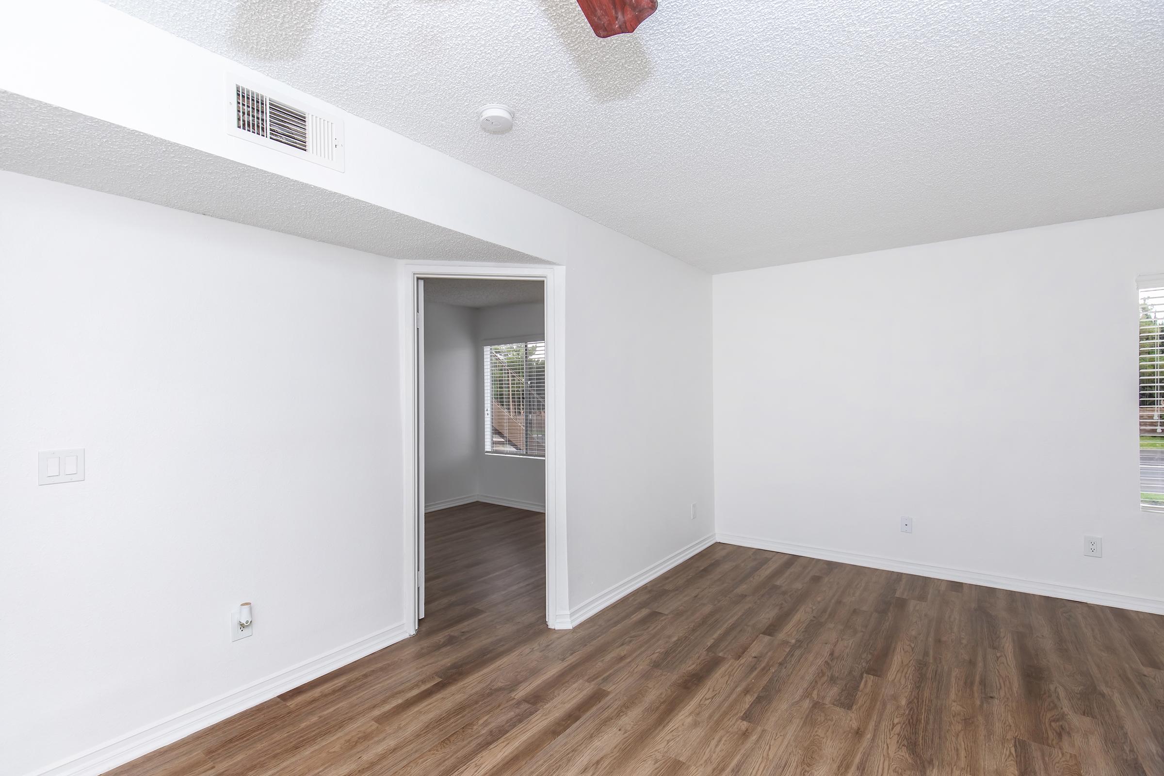 A spacious, empty room with light-colored walls and a textured ceiling. The flooring is wood-like, and there is a ceiling fan. To the left, an open doorway leads to another room with a window. Natural light streams in from the window, creating a bright and airy atmosphere.
