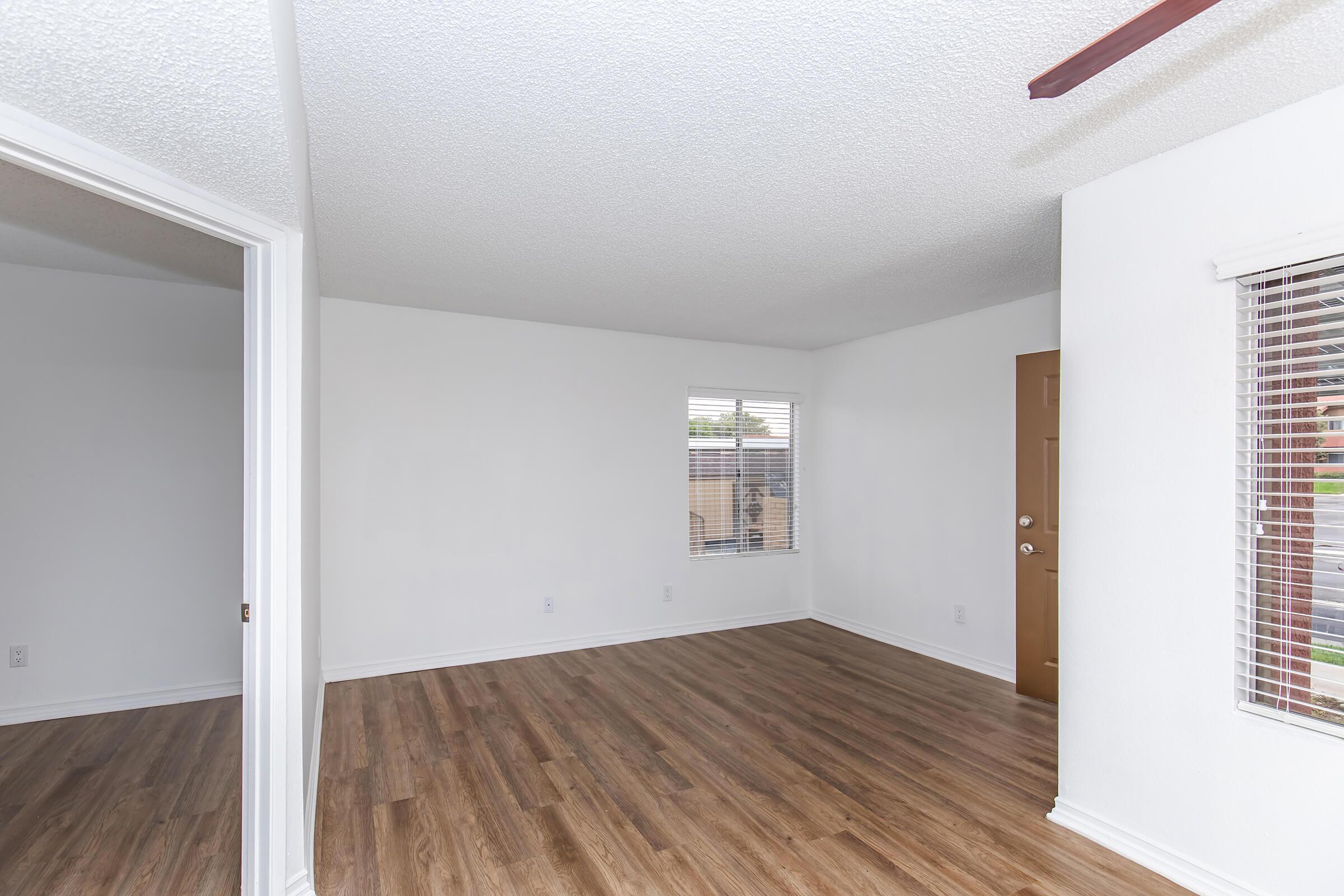 A bright, empty living room with light-colored walls and wooden flooring. A window with blinds lets in natural light, and a door is visible on the right. The room has a textured ceiling and a fan, creating a spacious and airy atmosphere.