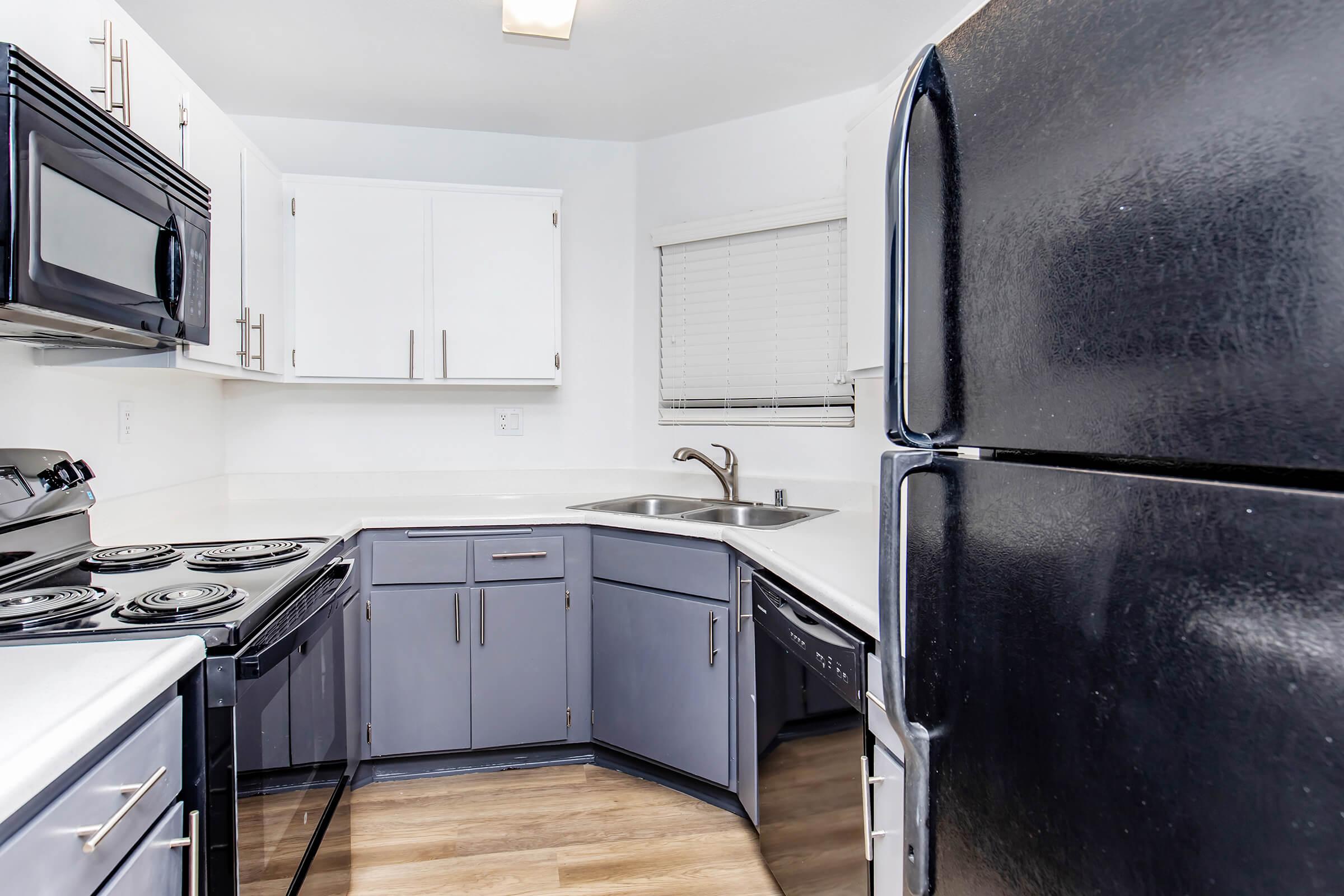 Modern kitchen featuring gray and white cabinetry, a black refrigerator, a black stove and microwave, and a stainless steel sink. The layout includes an L-shaped design with ample counter space. Light-colored flooring contrasts with the darker appliances, creating a bright and functional cooking area.