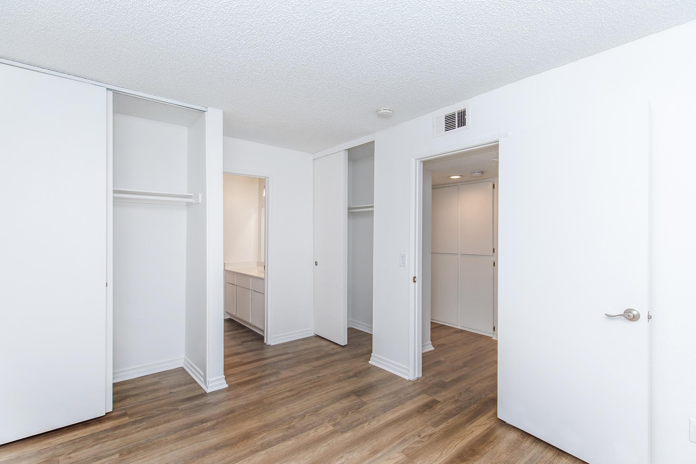 Interior of a modern room featuring light-colored walls, wooden flooring, and a minimalist design. There are two closet spaces with white sliding doors, a doorway leading to an adjoining room, and a bathroom vanity visible in the background. The lighting is bright, giving a spacious feel to the area.