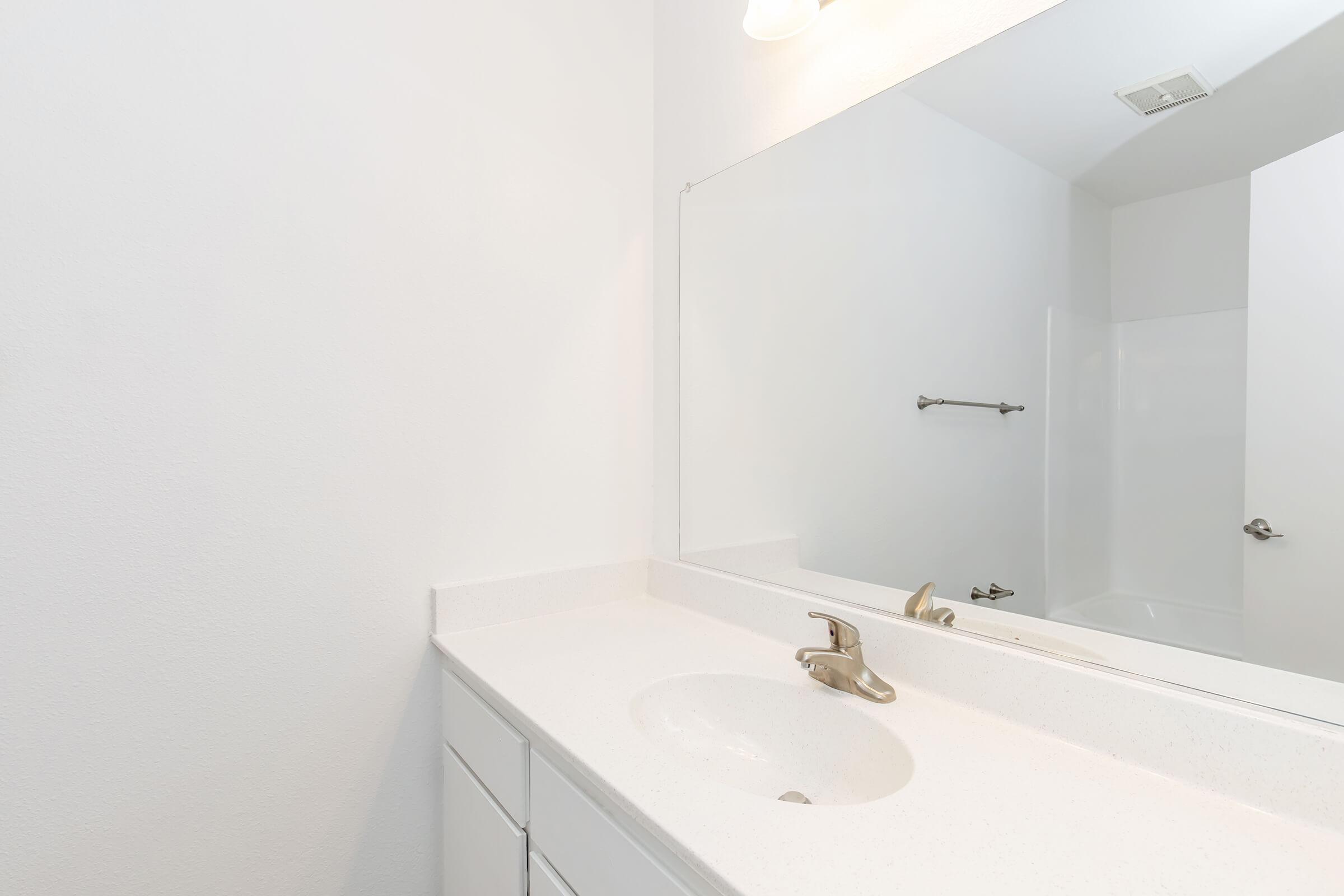 A clean, simple bathroom interior featuring a white countertop with an under-mounted sink and a bronze faucet. A large mirror is mounted above the sink, reflecting the bright, minimalist space. There is a towel bar on the wall and a glimpse of a bathtub in the background, emphasizing the modern design.