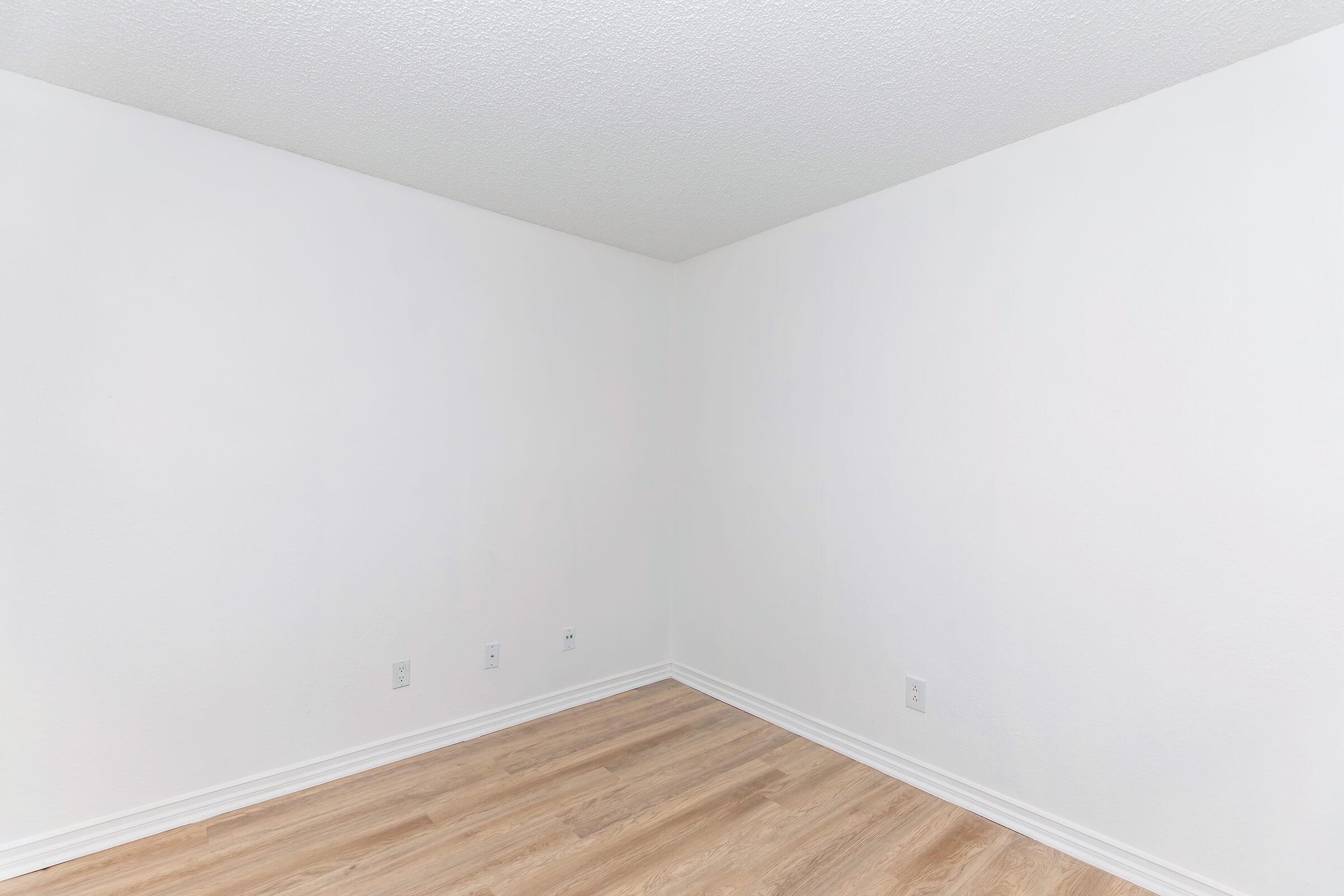 Empty room with white walls and light wooden flooring. One corner of the room is visible, showcasing a clean and minimalist space with no furniture or decorations. Natural light may be present, highlighting the simplicity of the interior.