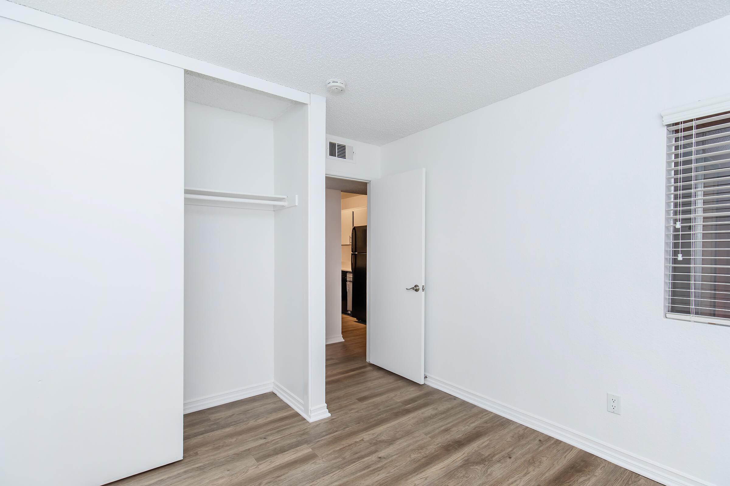 A bright, minimally furnished room featuring white walls and a wooden floor. On the left, there is a closet with a sliding door, and to the right is a partially open door leading to another area. Natural light filters in through a small window with blinds. The overall aesthetic is clean and modern.
