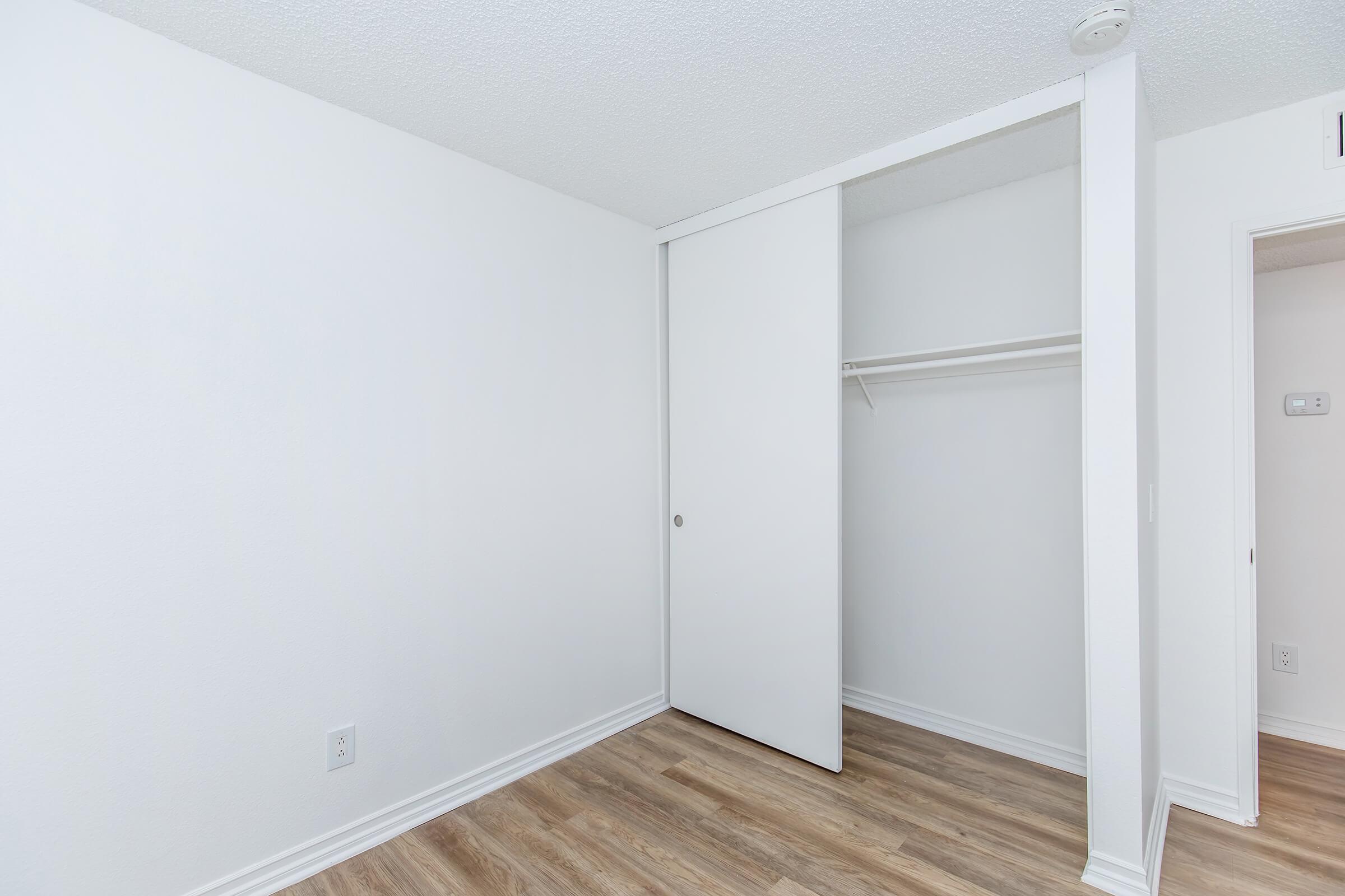 A bright, minimalistic room featuring white walls and a wooden floor. There is a sliding closet door on the left, with a single shelf visible inside. The space is uncluttered, ideal for storage or personal use, and has good natural light.