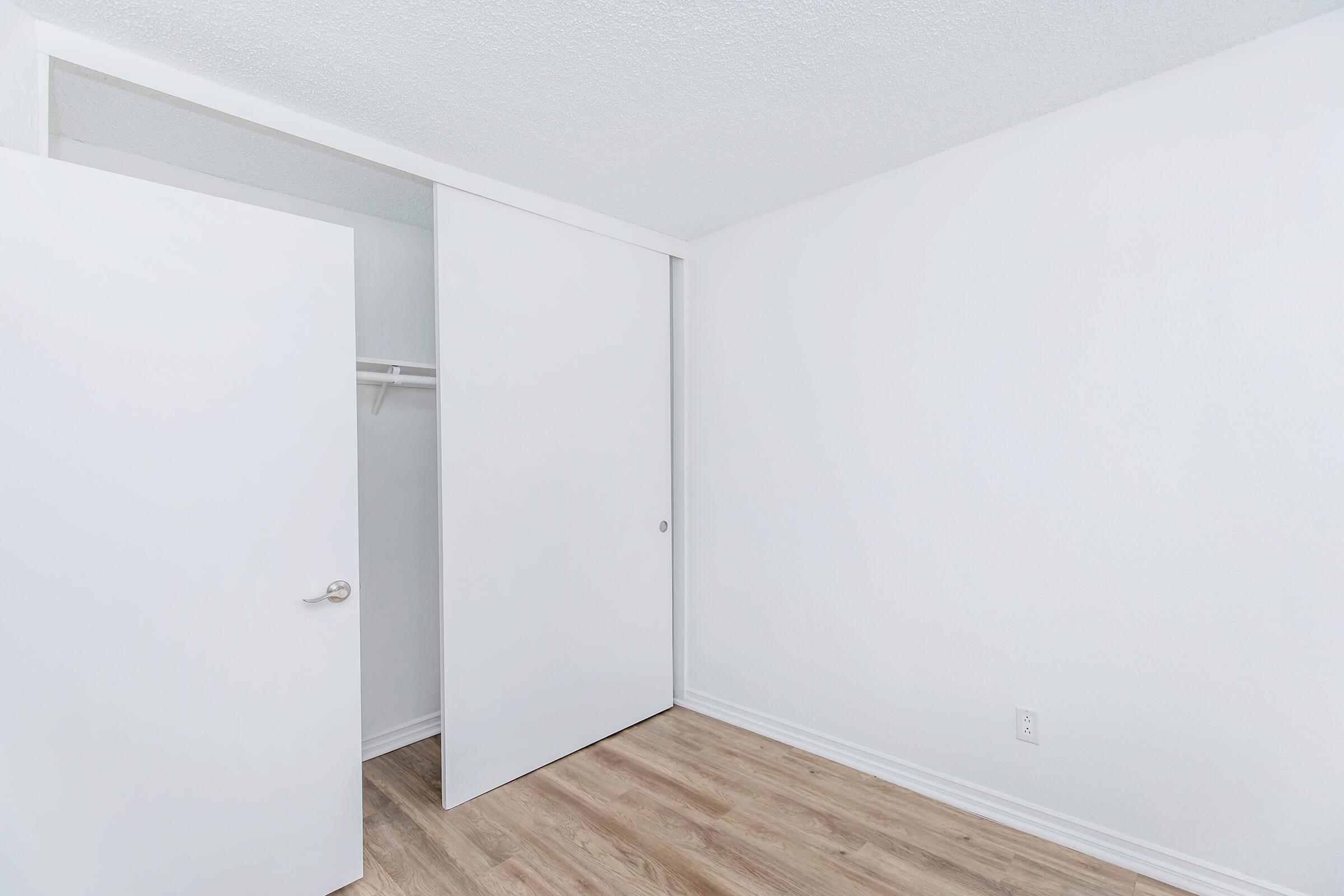 A well-lit, empty room with white walls and a light wood floor. There is a sliding door closet on one side, which is partially open, revealing a simple rod for hanging clothes. The overall appearance is clean and minimalistic, making it ideal for various uses or decoration.