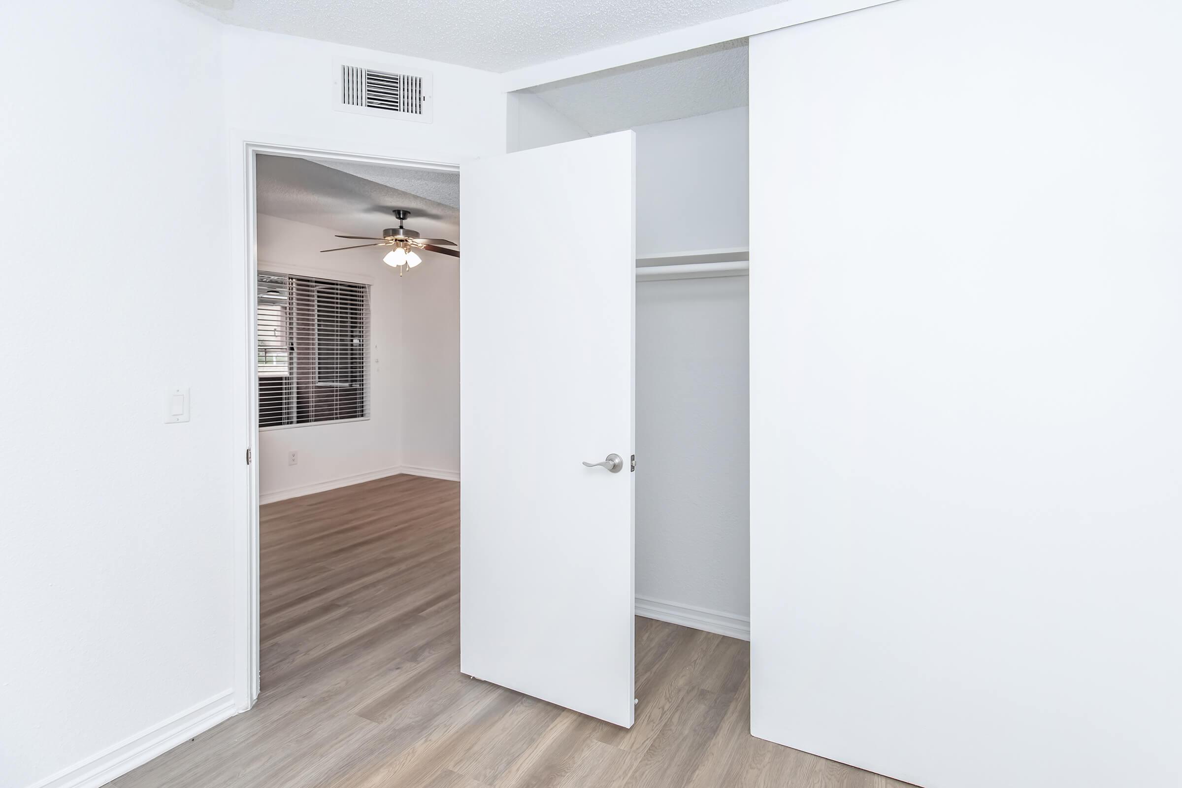 Interior view of a bright, modern room featuring an open closet with white doors. The floor is light-colored wood, and a ceiling fan is visible in the background. The walls are painted white, enhancing the spacious and clean aesthetic of the area. Natural light is suggested by the window on the far side.
