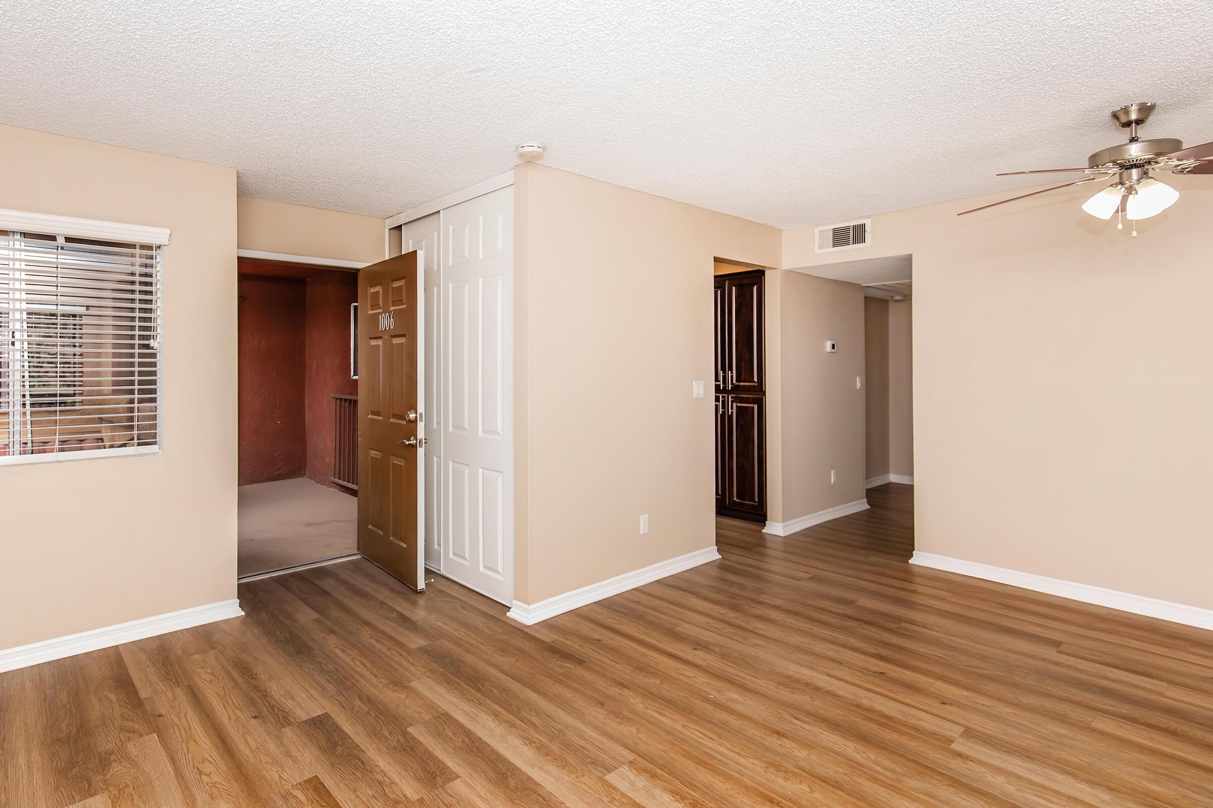 Interior view of an apartment featuring light-colored walls, a wooden door leading outside, and brown laminate flooring. A ceiling fan is visible, along with a large window with blinds. The space appears open and well-lit, showing two doorways leading to other areas of the home.