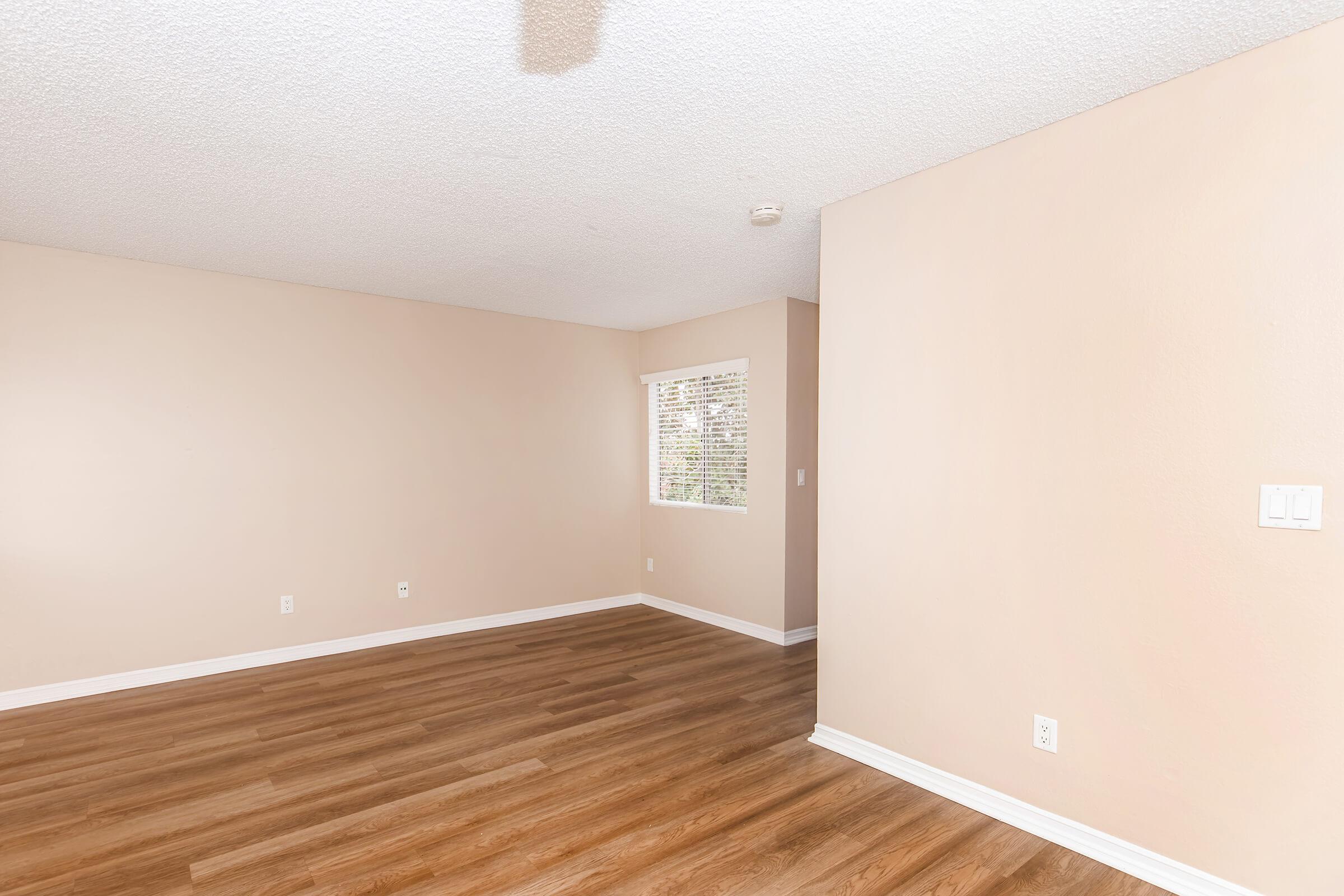 An empty room with light beige walls and a wooden floor. A small window with blinds is visible on one wall, allowing natural light to enter. The space has no furniture or decorations, creating a clean and minimalistic atmosphere.