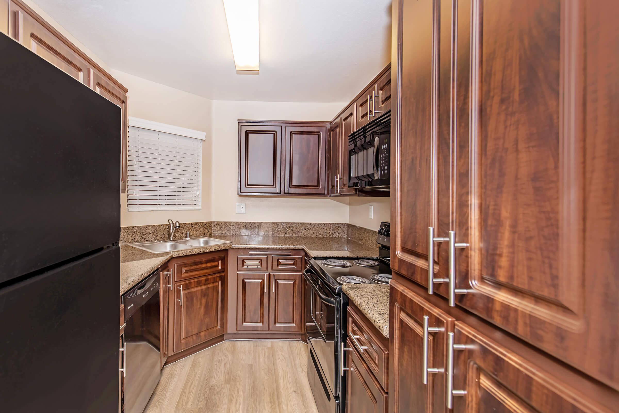 A modern kitchen featuring dark wood cabinetry, granite countertops, and stainless steel appliances. The layout includes a corner sink and a black refrigerator, with a window providing natural light. The overall design is sleek and functional.