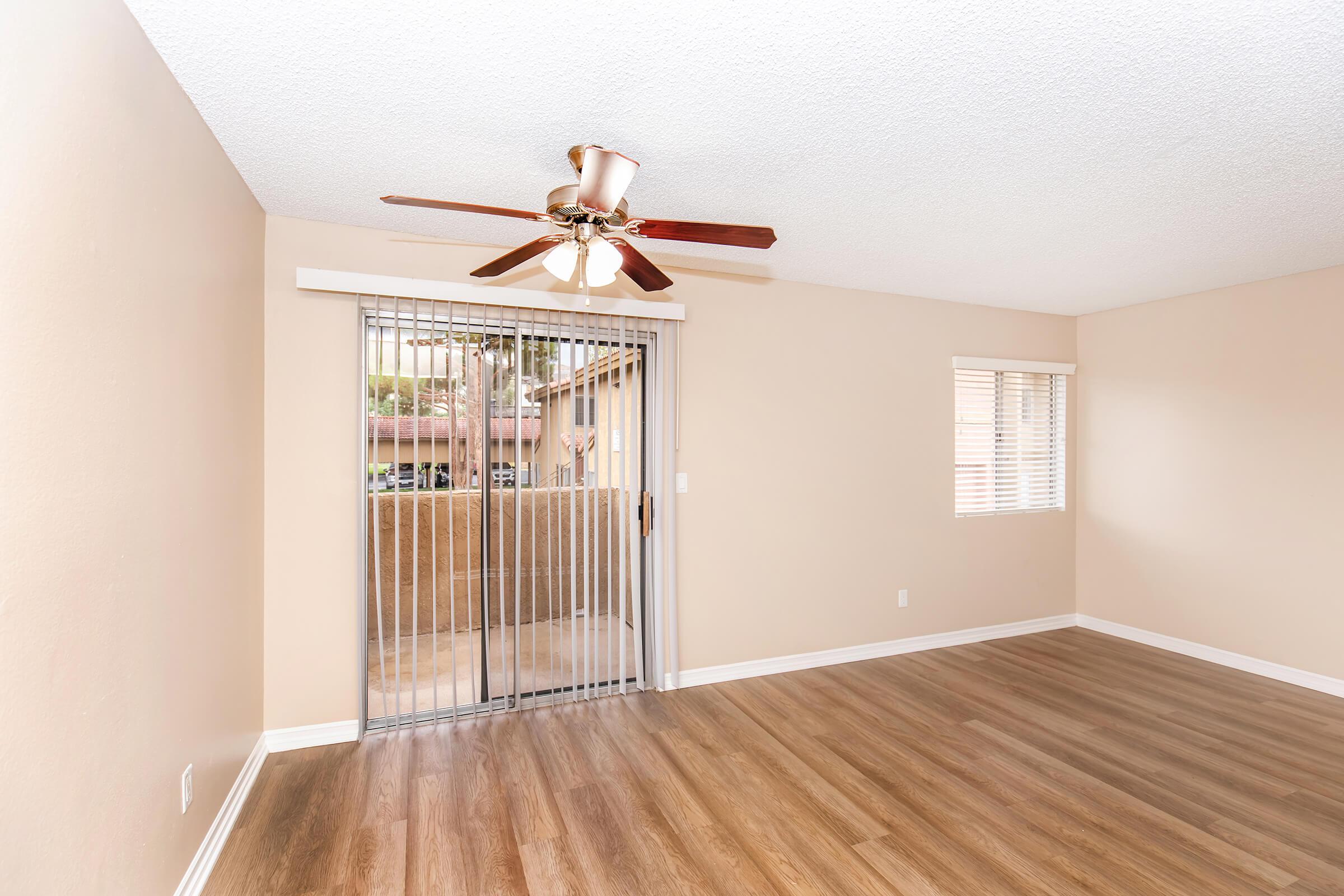 A spacious room with light brown walls and wooden flooring. A ceiling fan with three lights hangs from the ceiling. Large sliding glass doors with a metallic security screen lead to a balcony area. A smaller window with blinds is on the side, allowing natural light to enter.