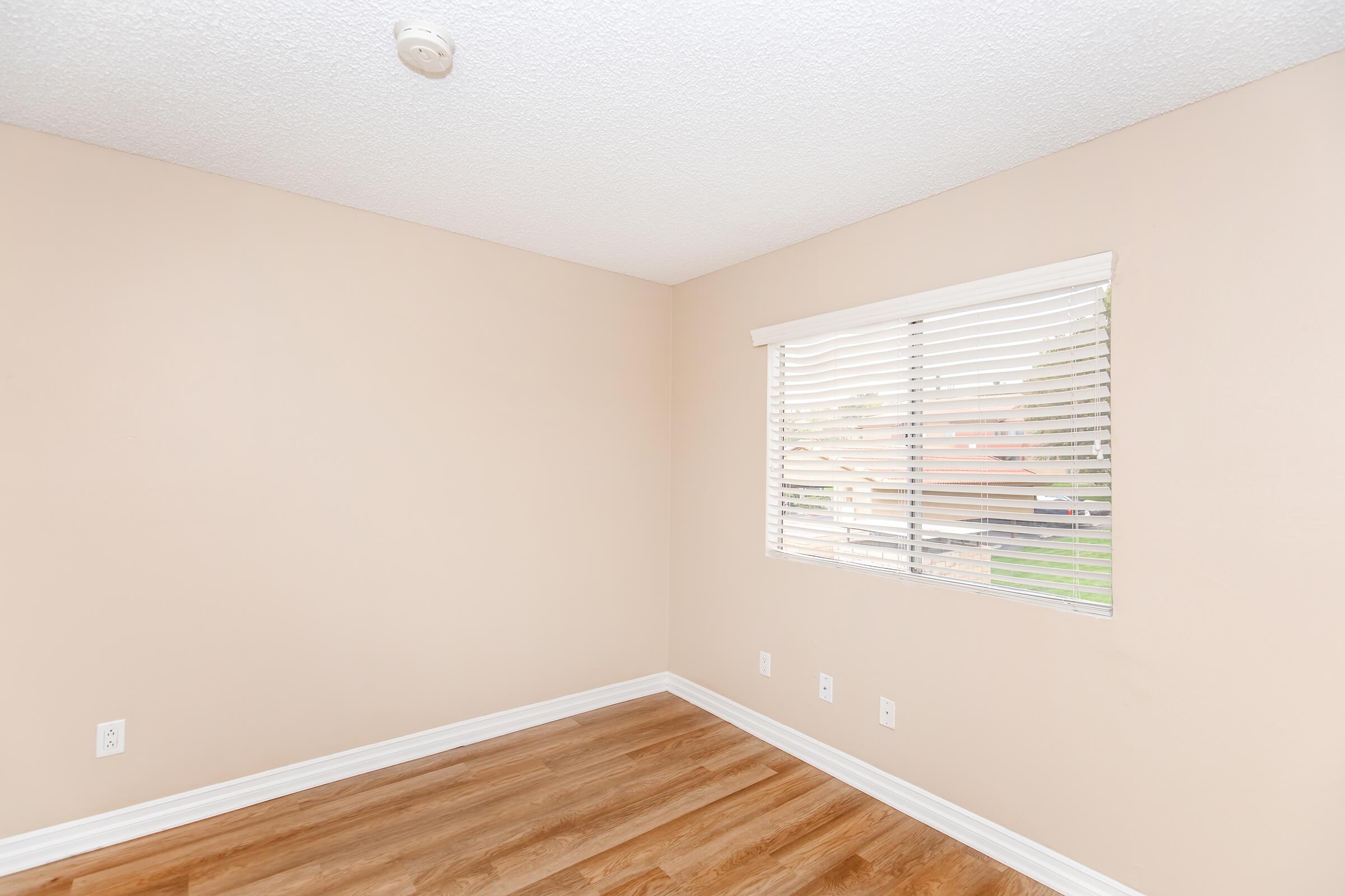 Empty room with light beige walls and hardwood flooring. A window with white blinds allows natural light to enter, offering a view of the outside. The ceiling features a white textured surface, and there are electrical outlets visible on the walls. The room is spacious and uncluttered.