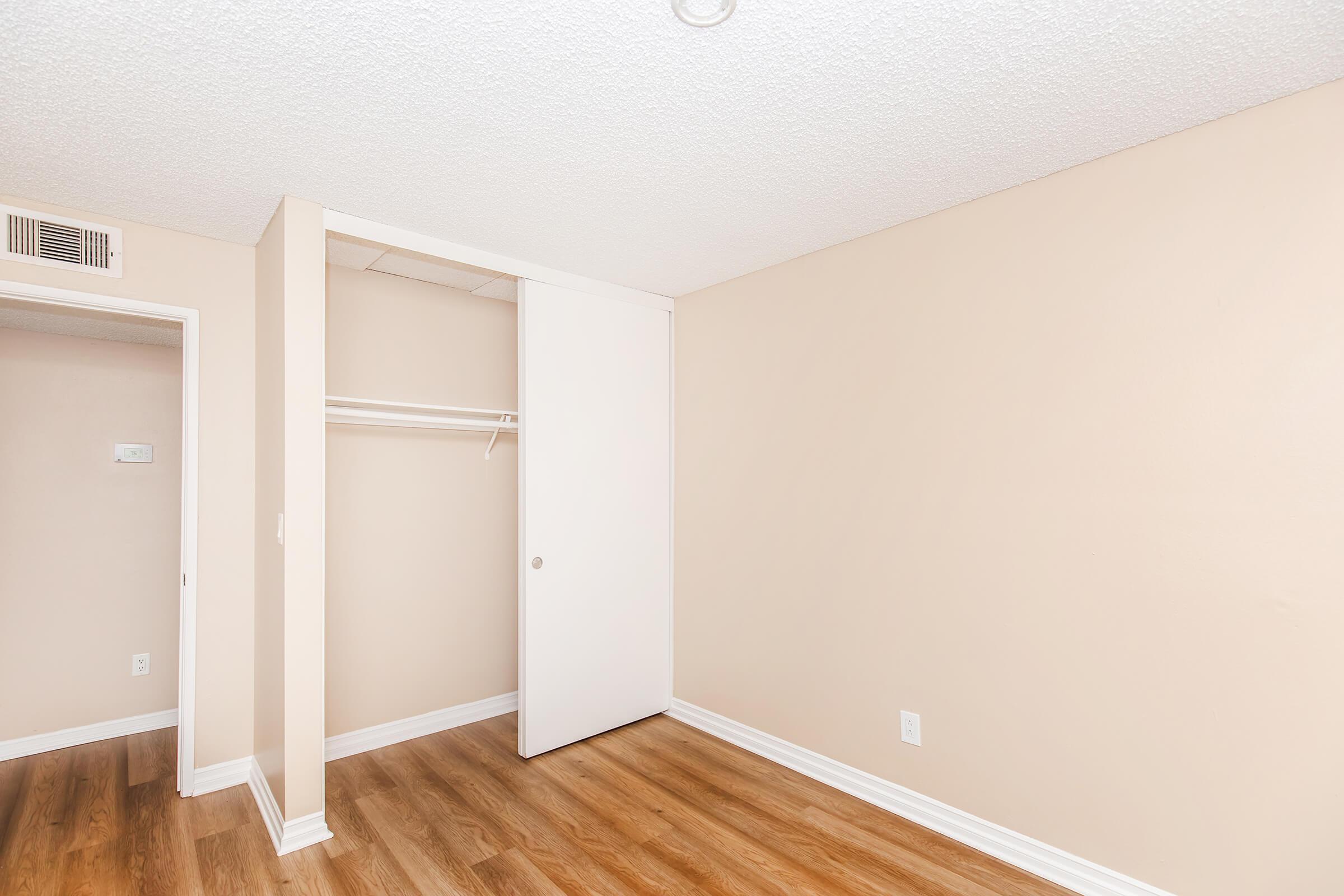 A small, empty room with light beige walls and wooden laminate flooring. There is a white sliding closet door on the left side and a wall with a small doorframe leading to another room. The space is well-lit and appears clean and freshly painted.