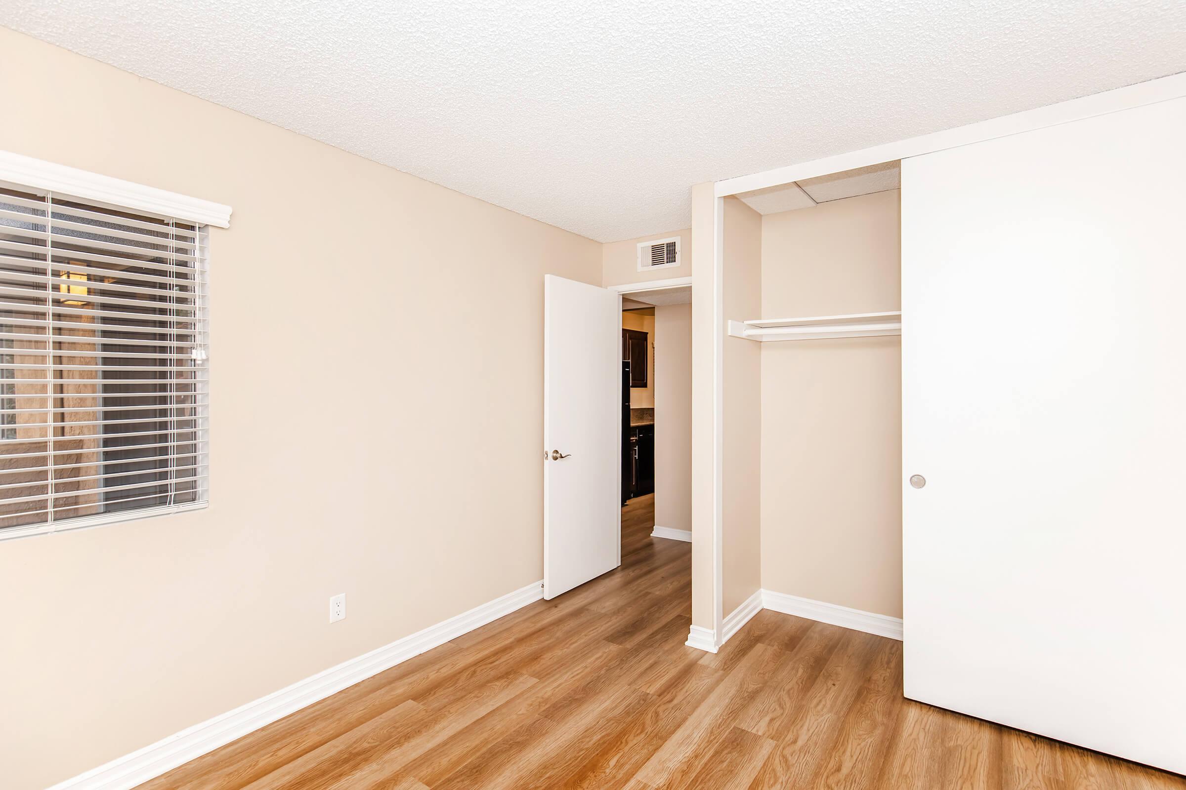 Empty room with beige walls, wood-like flooring, and a window with blinds on the left. A white closet door and a door leading to another room are visible. The space appears freshly painted and well-lit, creating a clean and inviting atmosphere.