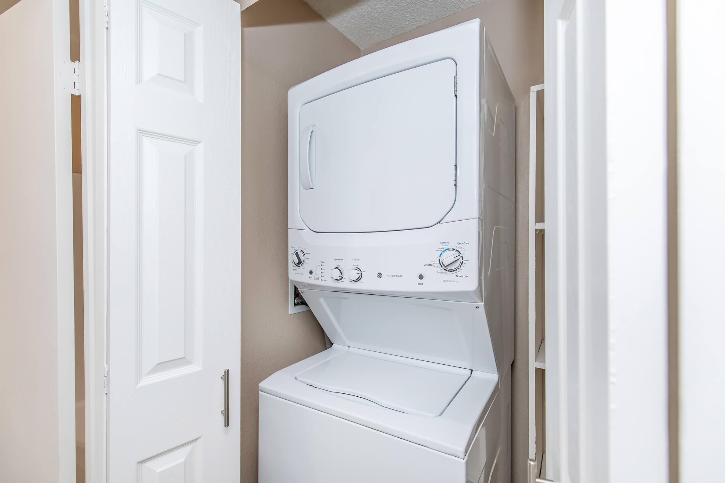 Stacked washer and dryer unit in a small laundry nook, flanked by white doors. The appliances are in a light-colored room with beige walls, and there is a shelf partially visible on the right. The washer is on the bottom, and the dryer is on top, both in a modern design.