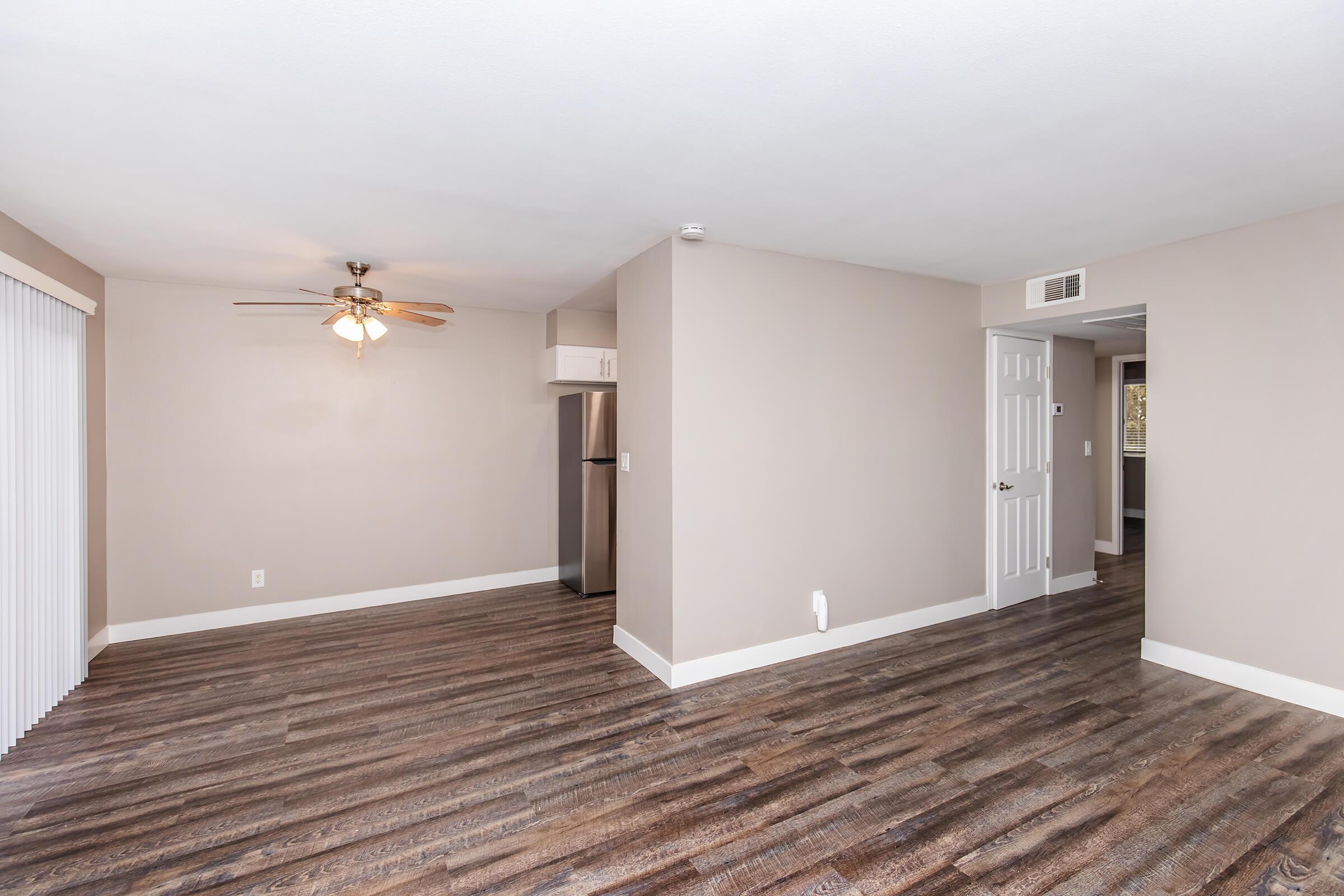 A spacious, vacant living area featuring light-colored walls, a ceiling fan, and wooden flooring. The room includes a doorway leading to another area and a sliding door with vertical blinds. The layout is open, allowing natural light to brighten the interior space.