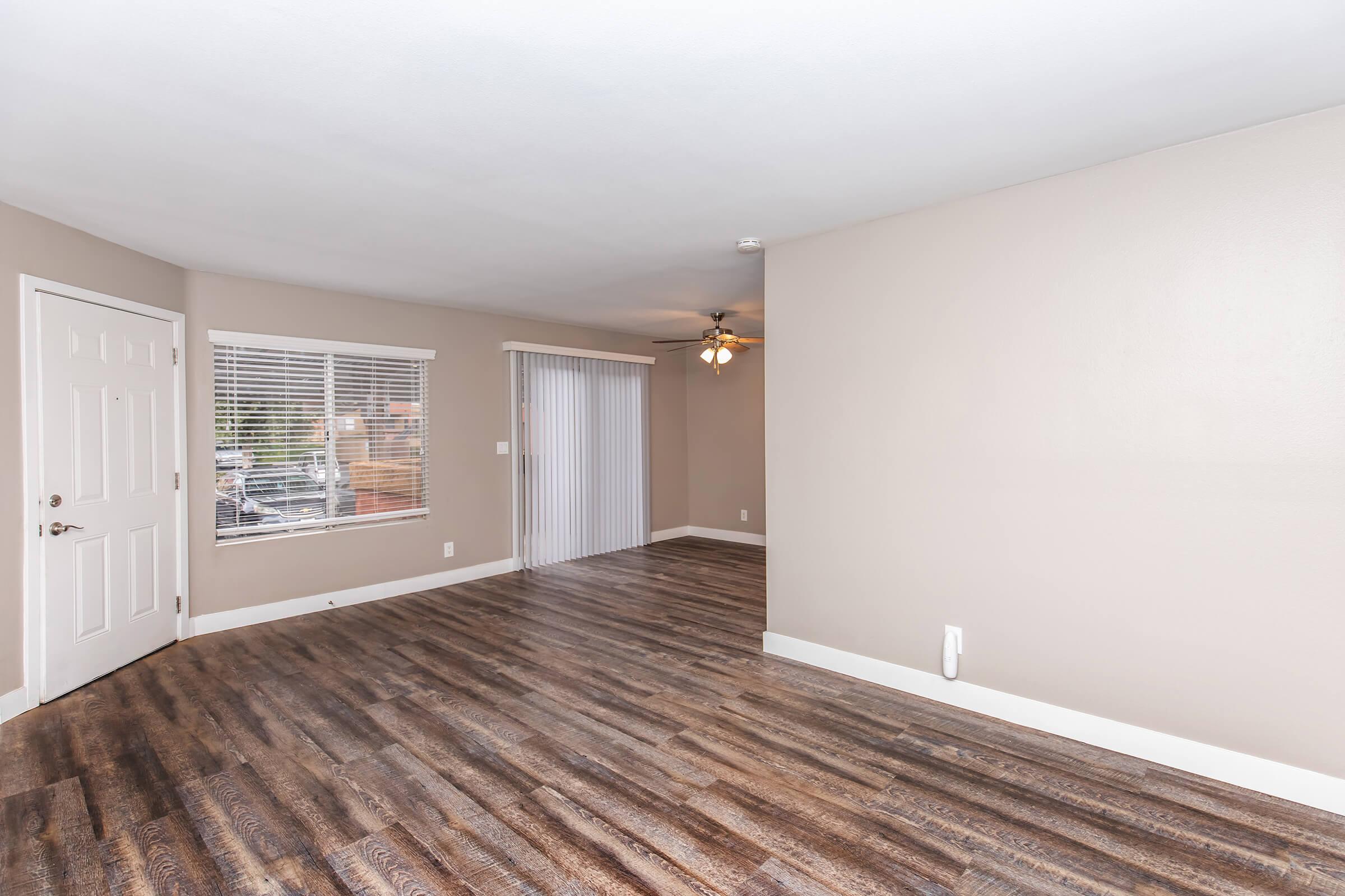 Interior of a room with light brown walls and hardwood flooring. Features include a white door, a window with blinds, and a sliding glass door leading to an outdoor area. A ceiling fan is visible, providing a bright atmosphere in the space. The room appears spacious and well-lit.