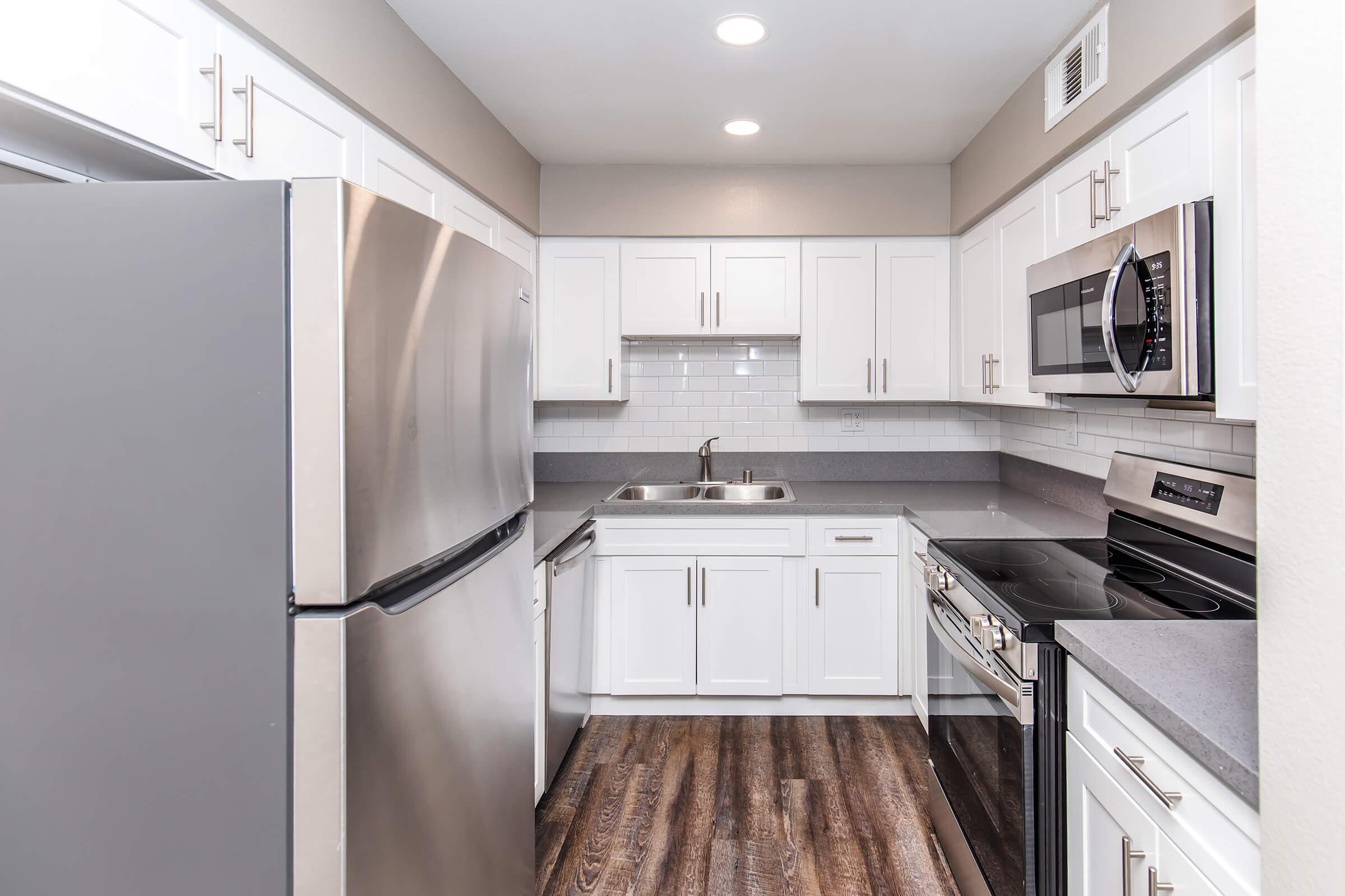 Modern kitchen featuring stainless steel appliances, including a refrigerator, microwave, and oven, with white cabinetry and grey countertops. The space has a tiled backsplash and is well-lit with overhead lights, complemented by a dark wood floor.