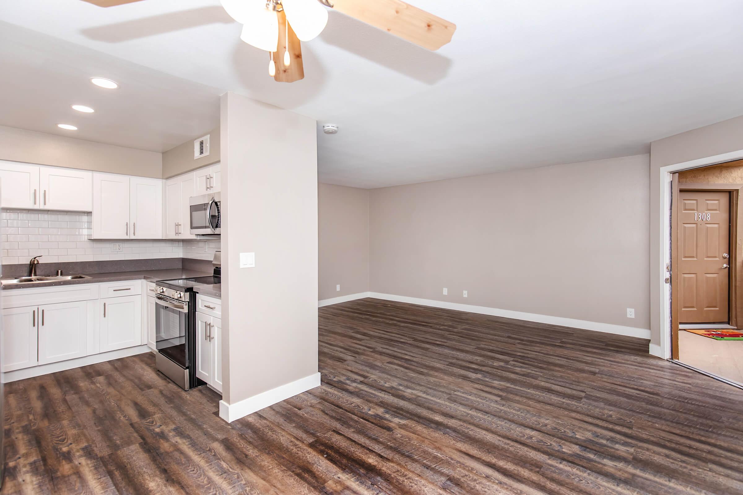 Interior view of a modern, empty living space featuring a kitchen with white cabinetry, stainless steel appliances, and a ceiling fan. The open floor plan leads to a light-colored wall and a door that opens to the outside. The flooring is dark wood, enhancing the spacious feel of the room.