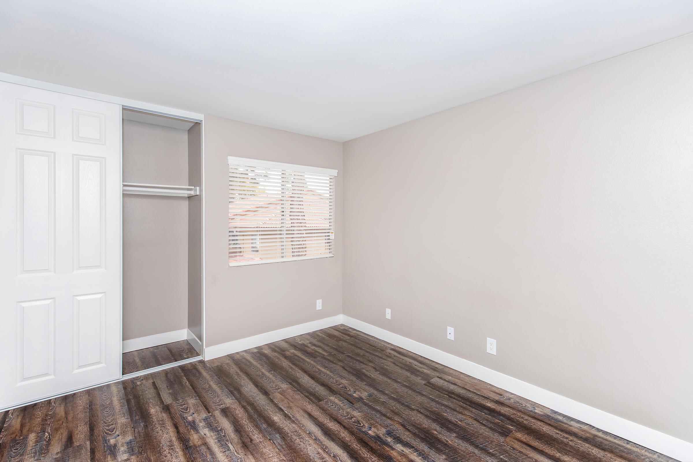 A spacious, empty room with light beige walls, a large window with white blinds, and a closet with sliding doors. The floor is dark wood laminate, and there are no furnishings or decorations present.
