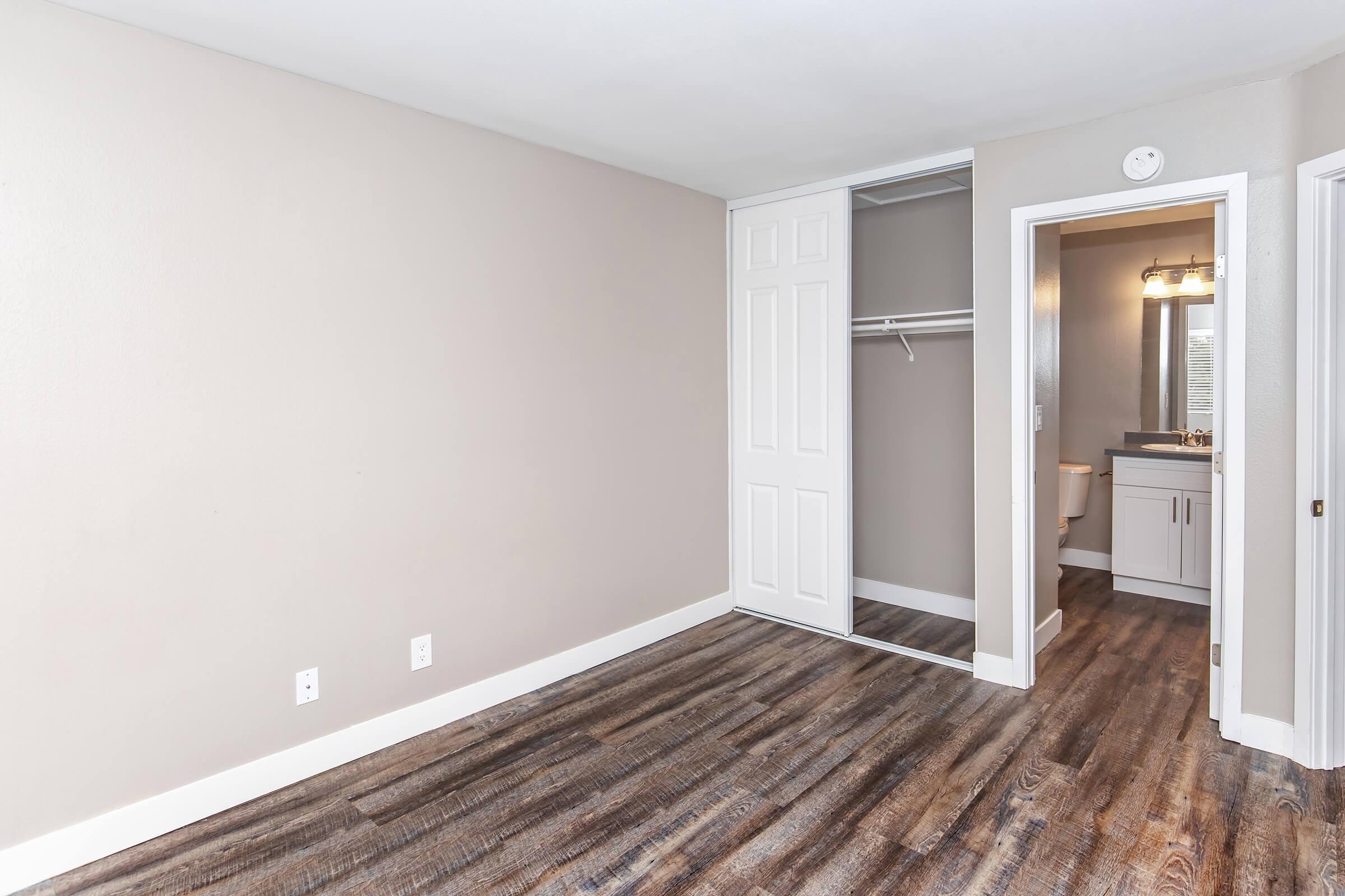 A small, empty bedroom with neutral walls, featuring a closet with sliding doors. The floor is covered in dark wood-like laminate. A doorway leads to a bathroom visible in the background, which is well-lit by overhead lighting. Natural light may enter through a window not shown in the image.