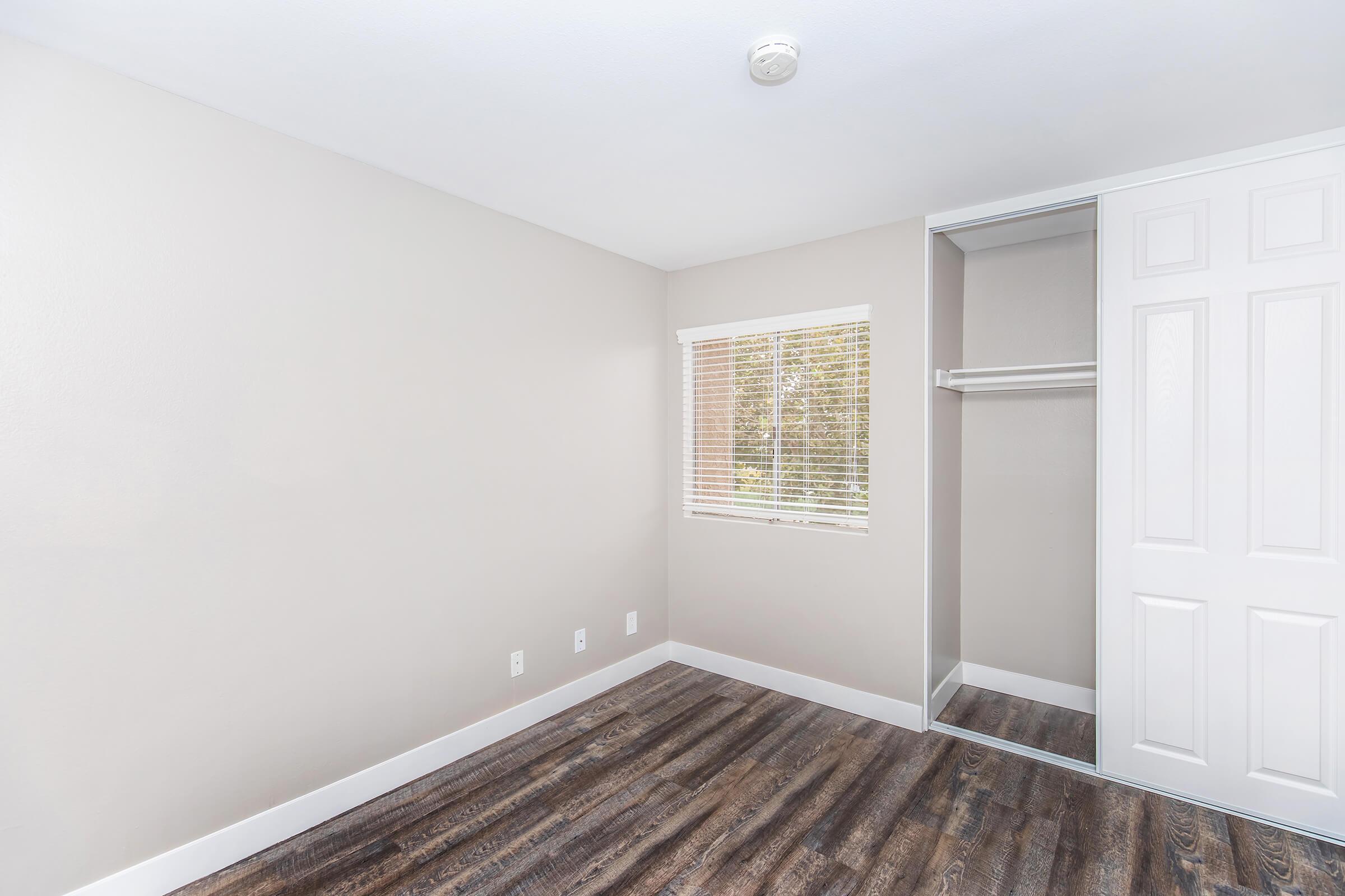 Empty bedroom with light-colored walls and a window allowing natural light. Features include a closet with sliding doors, hardwood flooring, and minimal furnishings, creating a spacious and tidy appearance.
