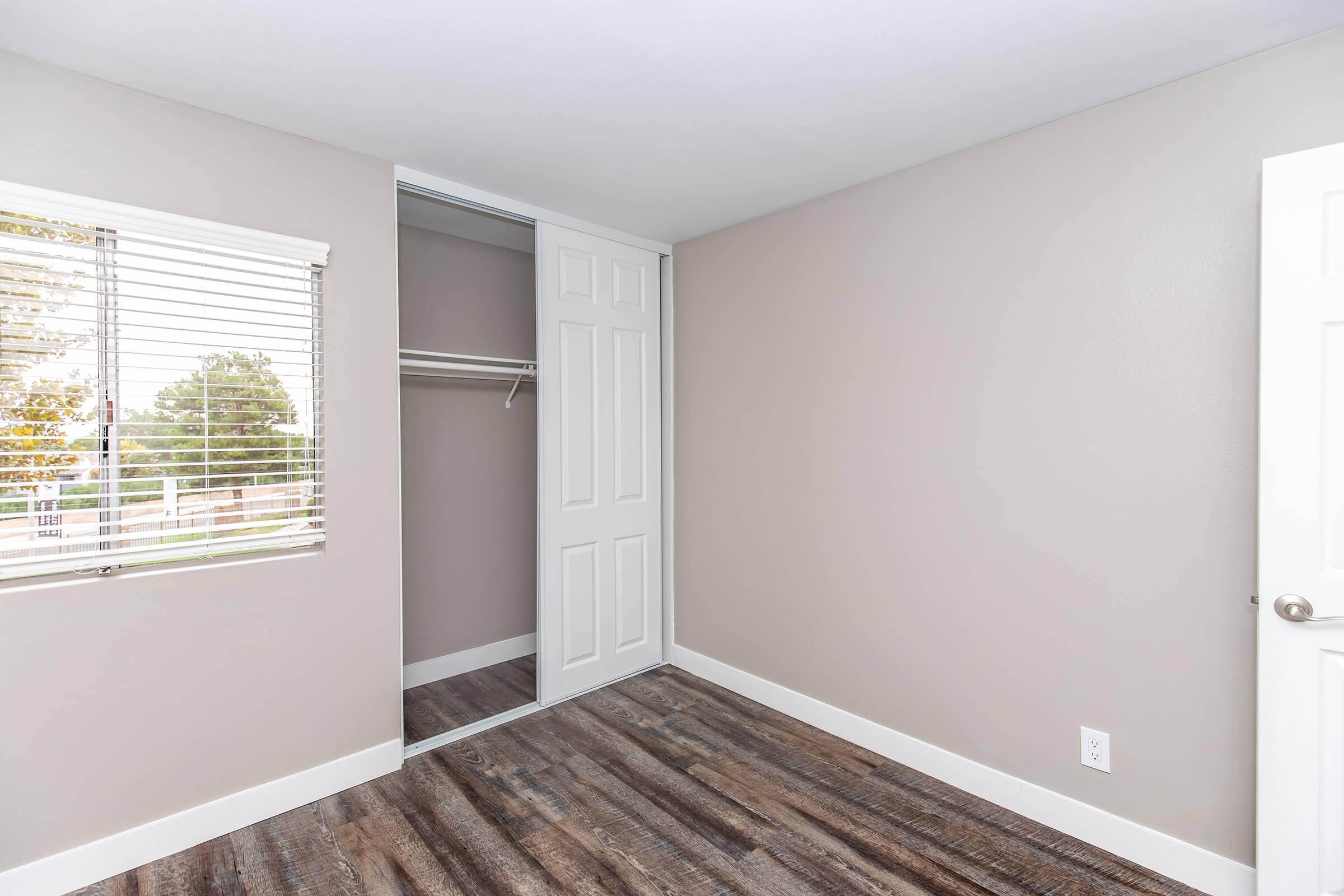 A well-lit, empty bedroom featuring light beige walls, a window with blinds, and a closet with sliding doors. The floor is made of wood-like planks, providing a clean and modern look.