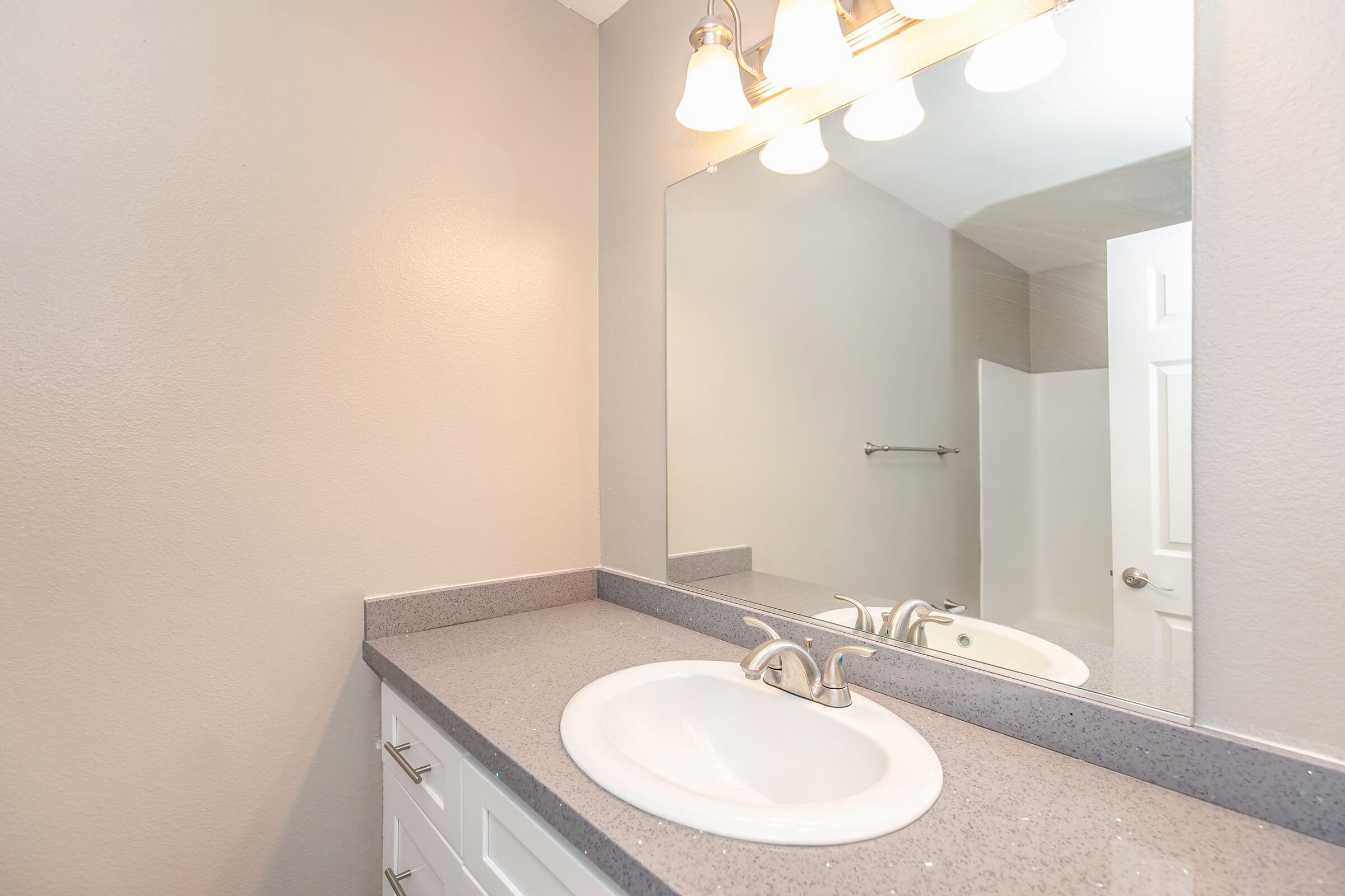 A clean, modern bathroom featuring a white sink with a chrome faucet, a light gray countertop, and a large mirror above. The walls are painted a light gray, and there is a towel bar mounted on the wall. Soft lighting is provided by three light fixtures above the mirror.