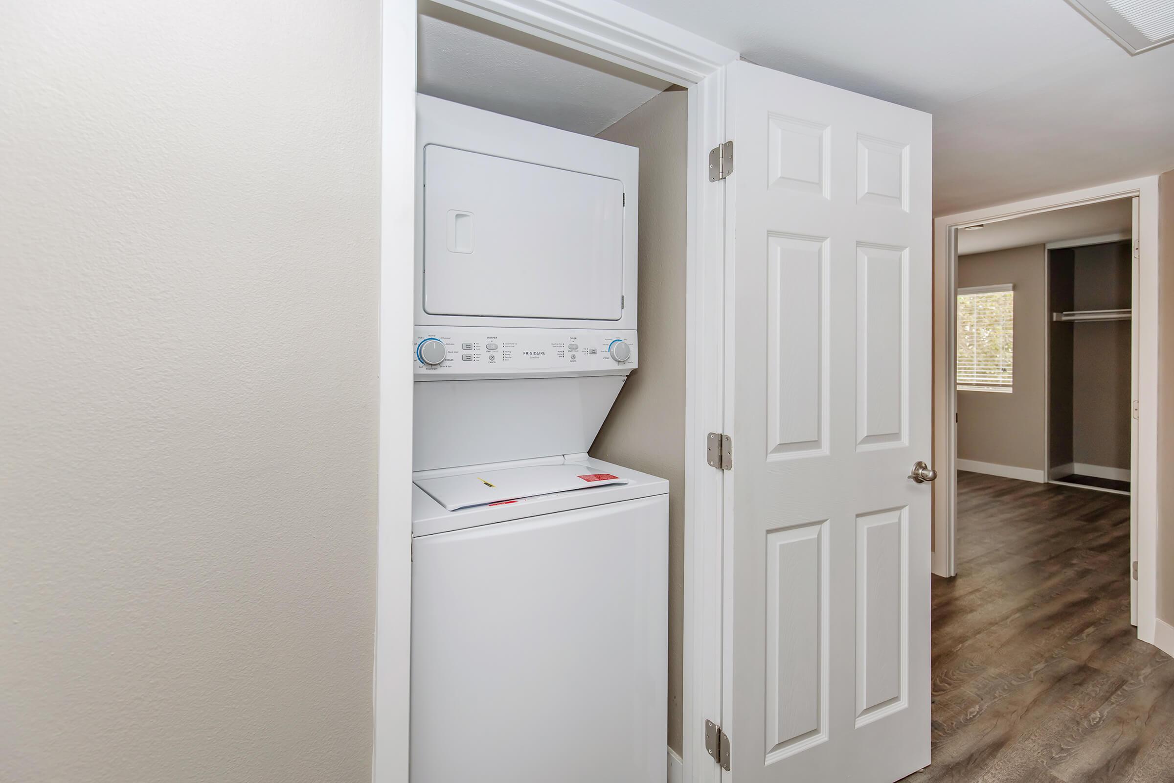 A small laundry area featuring a stacked washer and dryer inside a closet. A door is partially open, revealing the appliances. The surrounding walls are painted in a light color, and there is a room with wooden flooring visible through another doorway.