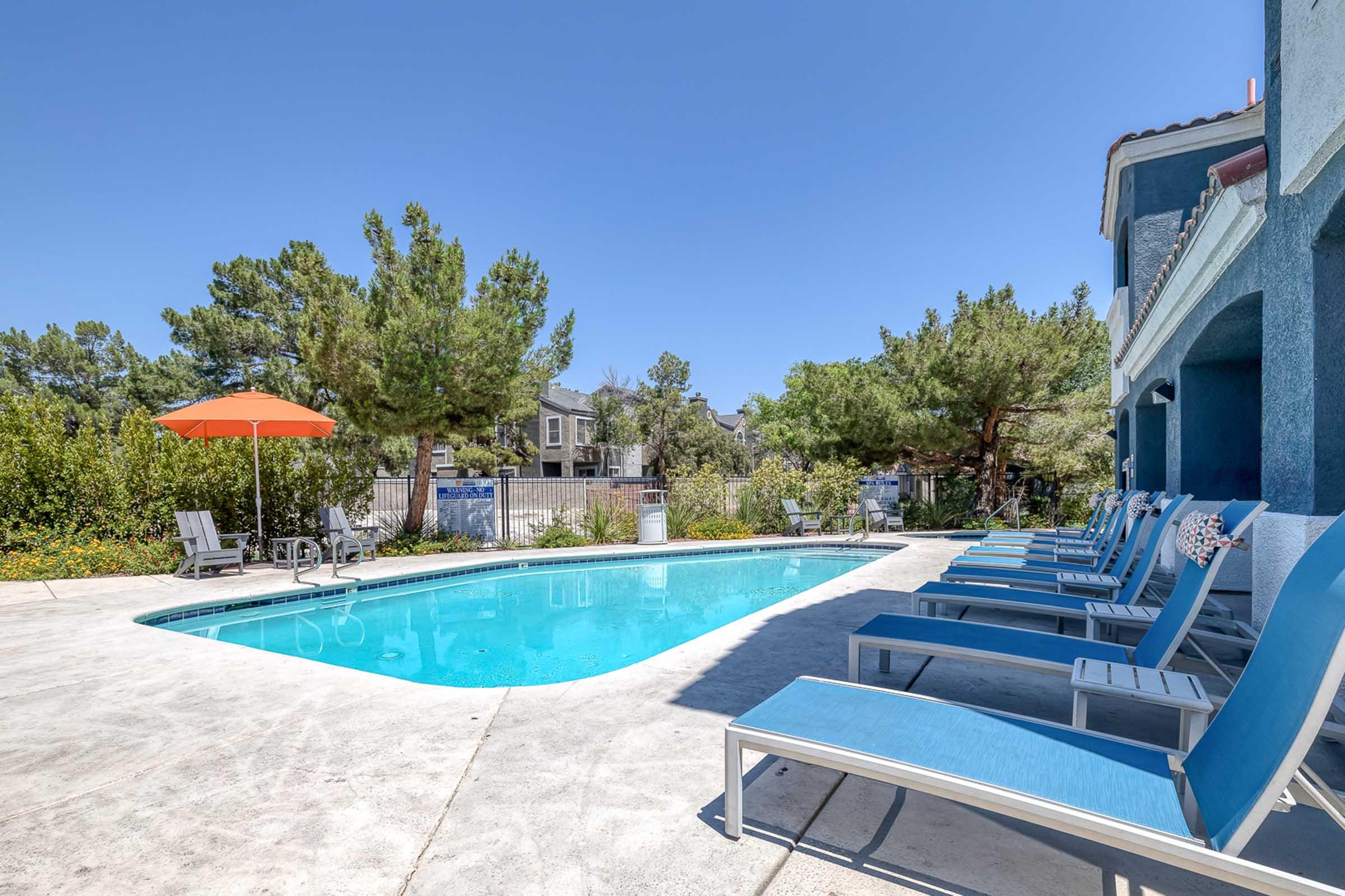 A tranquil swimming pool area featuring a clear blue pool surrounded by comfortable lounge chairs. An orange patio umbrella provides shade, while lush greenery and residential structures are visible in the background under a bright blue sky.