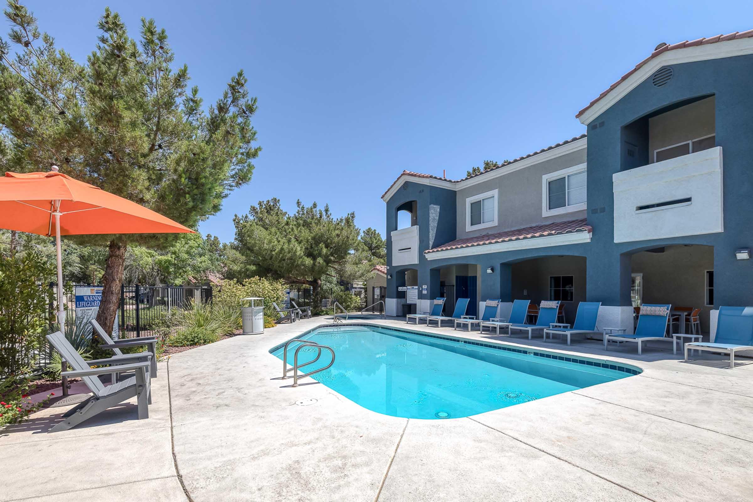 A clear blue pool surrounded by lounge chairs and a bright orange umbrella, set in a sunny outdoor area with trees and a two-story building in the background. The scene conveys a relaxing and inviting atmosphere perfect for leisure.