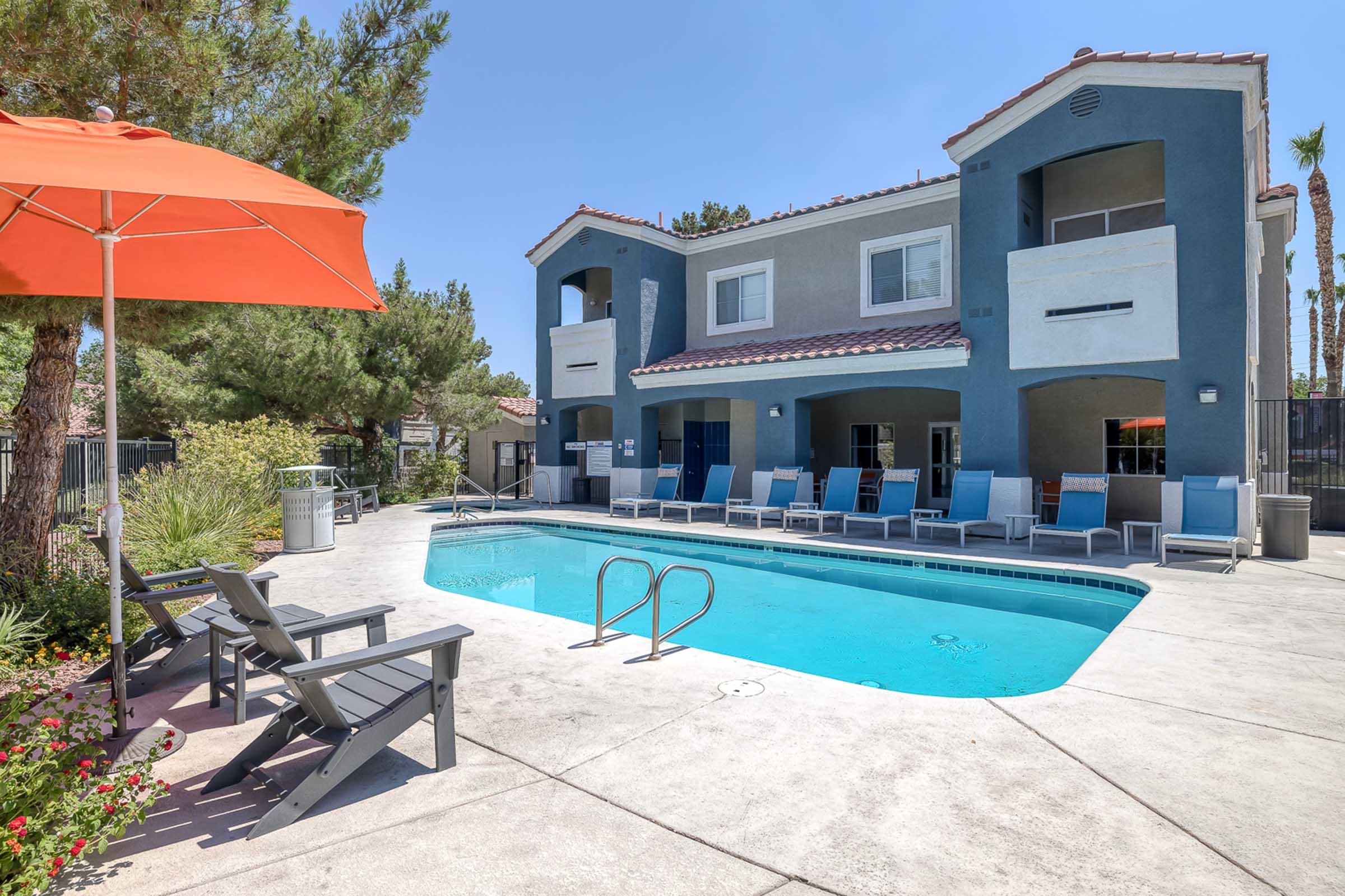 View of a sunny outdoor pool area at an apartment complex, featuring a clear blue pool, lounge chairs, and a vibrant orange umbrella. Surrounding greenery adds a touch of nature, while the two-story building in the background completes the inviting atmosphere.