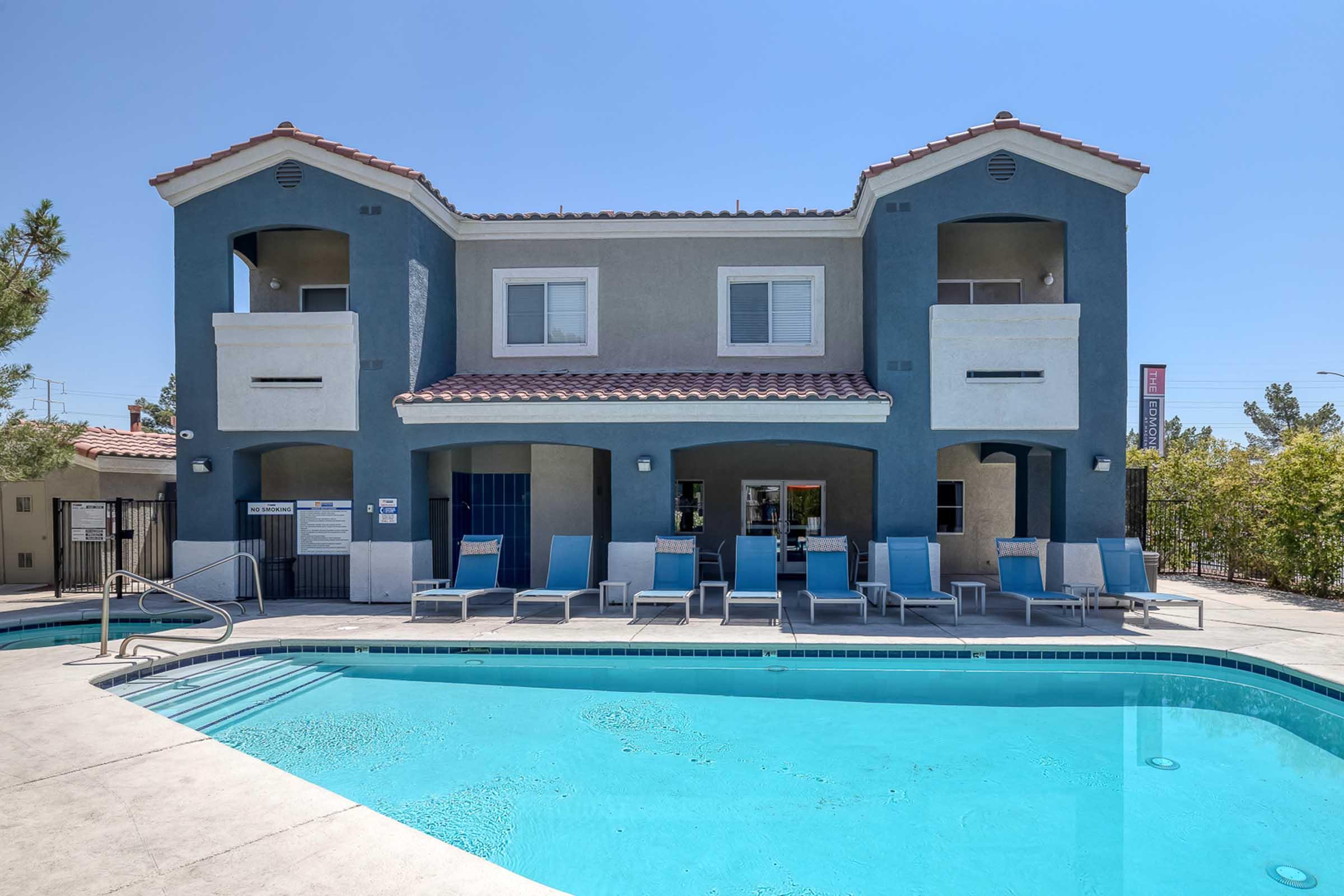 View of a two-story, blue and gray building with a pool in the foreground. The building features balconies, several lounge chairs by the pool, and a sunny sky. Surrounding landscaping adds greenery to the scene, creating a relaxing atmosphere.
