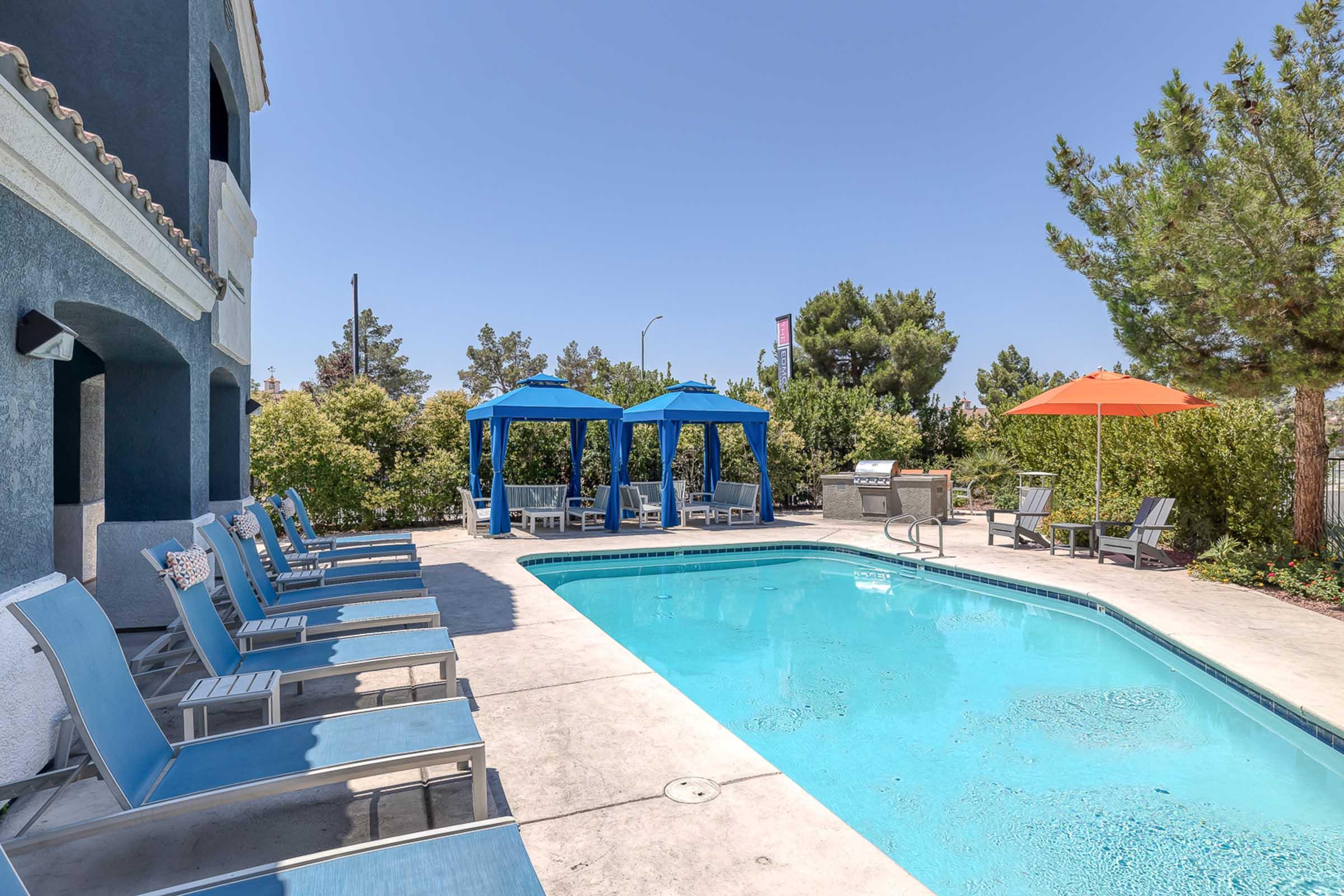 View of a sunny outdoor pool area at an apartment complex, featuring a clear blue pool, lounge chairs, and a vibrant orange umbrella.