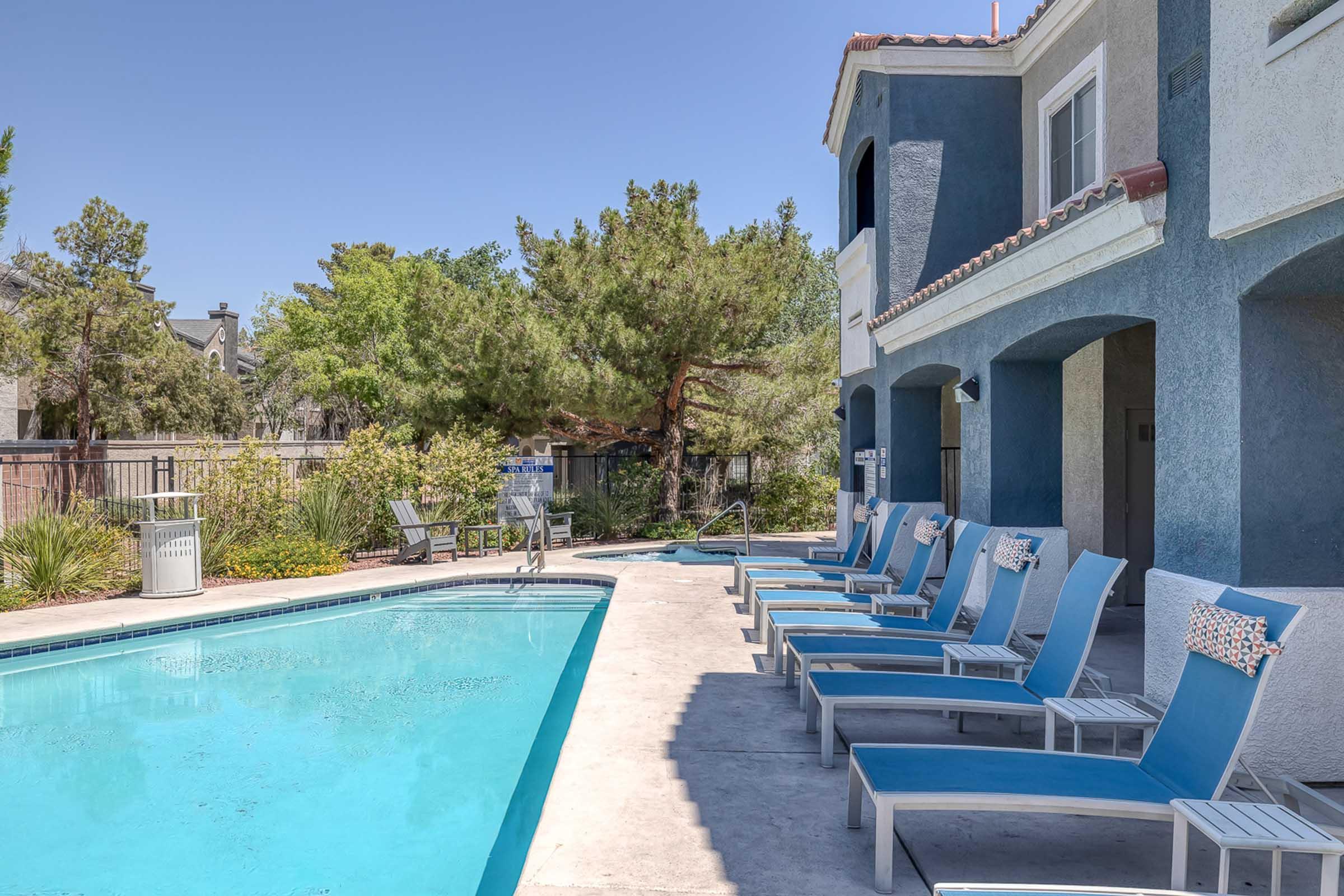 A tranquil pool area featuring a swimming pool surrounded by lounge chairs. Lush greenery and trees provide a natural backdrop. The two-story building nearby has a light-colored exterior and arched windows, suggesting a welcoming residential atmosphere. Clear skies overhead complete the serene setting.