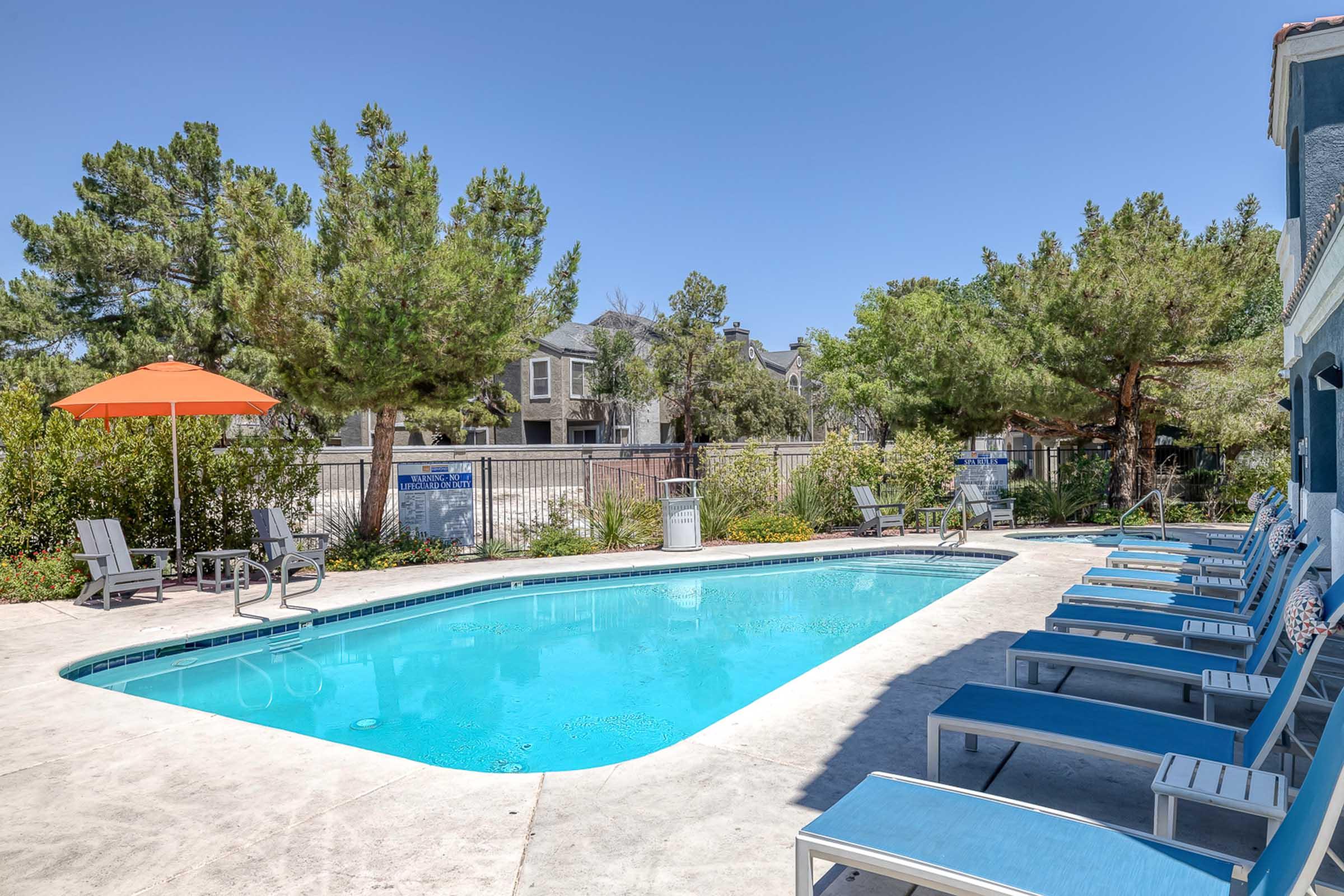 A clear swimming pool surrounded by lounge chairs and a bright orange umbrella. Green trees line the perimeter, and a fence can be seen in the background, along with some buildings peeking through the foliage. The scene is sunny and inviting, suggesting a relaxing outdoor space.