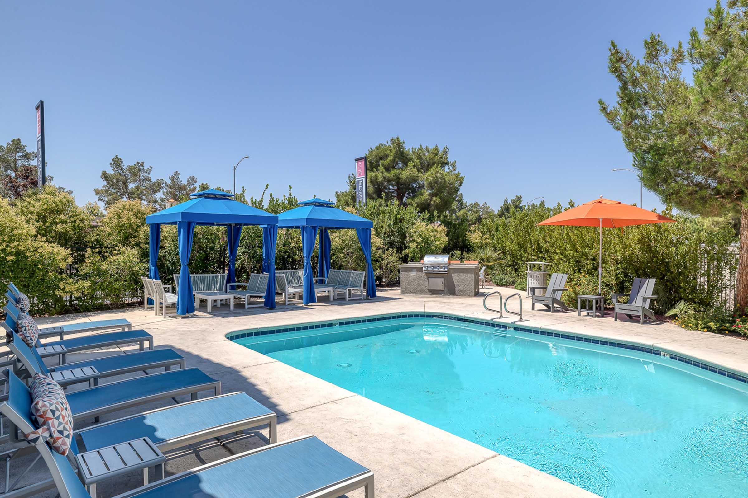 A sunny pool area featuring a clear blue swimming pool surrounded by lounge chairs. There are two blue cabanas and an orange umbrella providing shade, with a barbecue grill nearby and lush greenery in the background. Ideal setting for relaxation and outdoor gatherings.