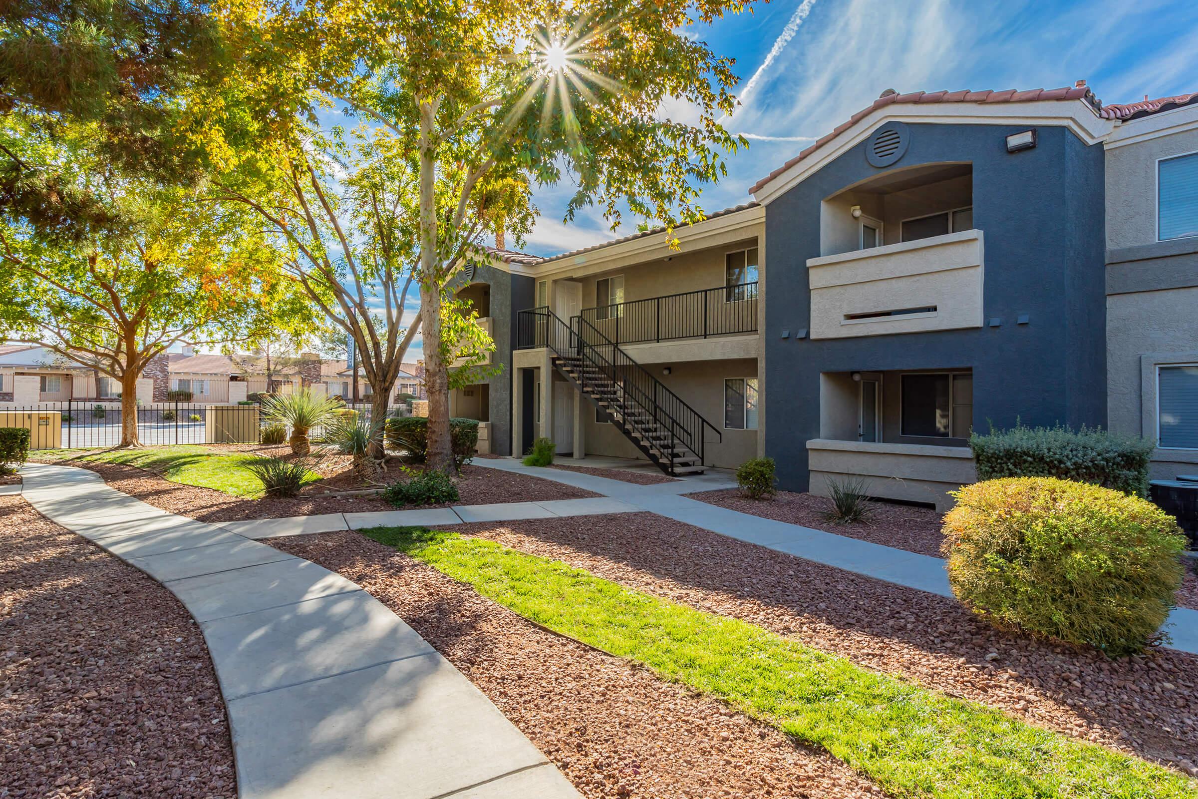 A well-maintained apartment complex exterior featuring a walkway bordered by grass and shrubs. There are two buildings with balconies, surrounded by trees and a clear blue sky. Sunlight shines brightly from behind the trees, creating a warm and inviting atmosphere.