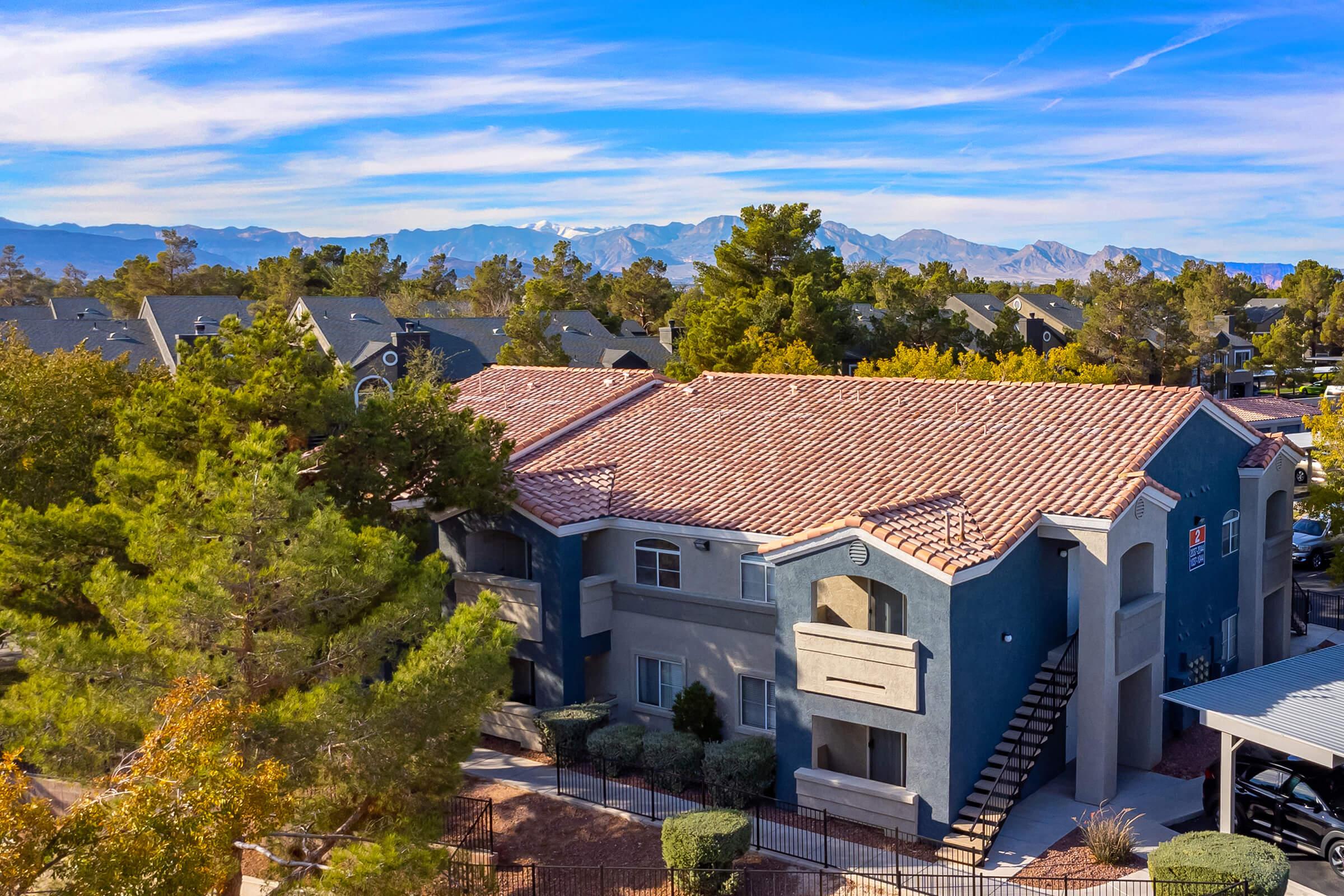 Aerial view of a modern apartment building with a terracotta tile roof, surrounded by trees and shrubs. In the background, mountains are visible under a clear blue sky with wispy clouds. The building has a staircase leading to upper units and features a blend of blue and gray colors.