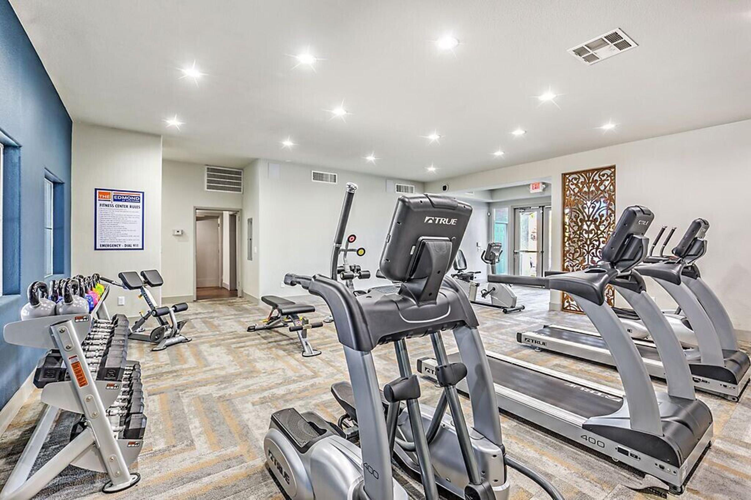 A modern fitness center featuring various exercise equipment, including treadmills, elliptical machines, and weights. The space is well-lit with recessed lighting and includes a decorative wall design. Large windows let in natural light, creating an inviting atmosphere for workouts.