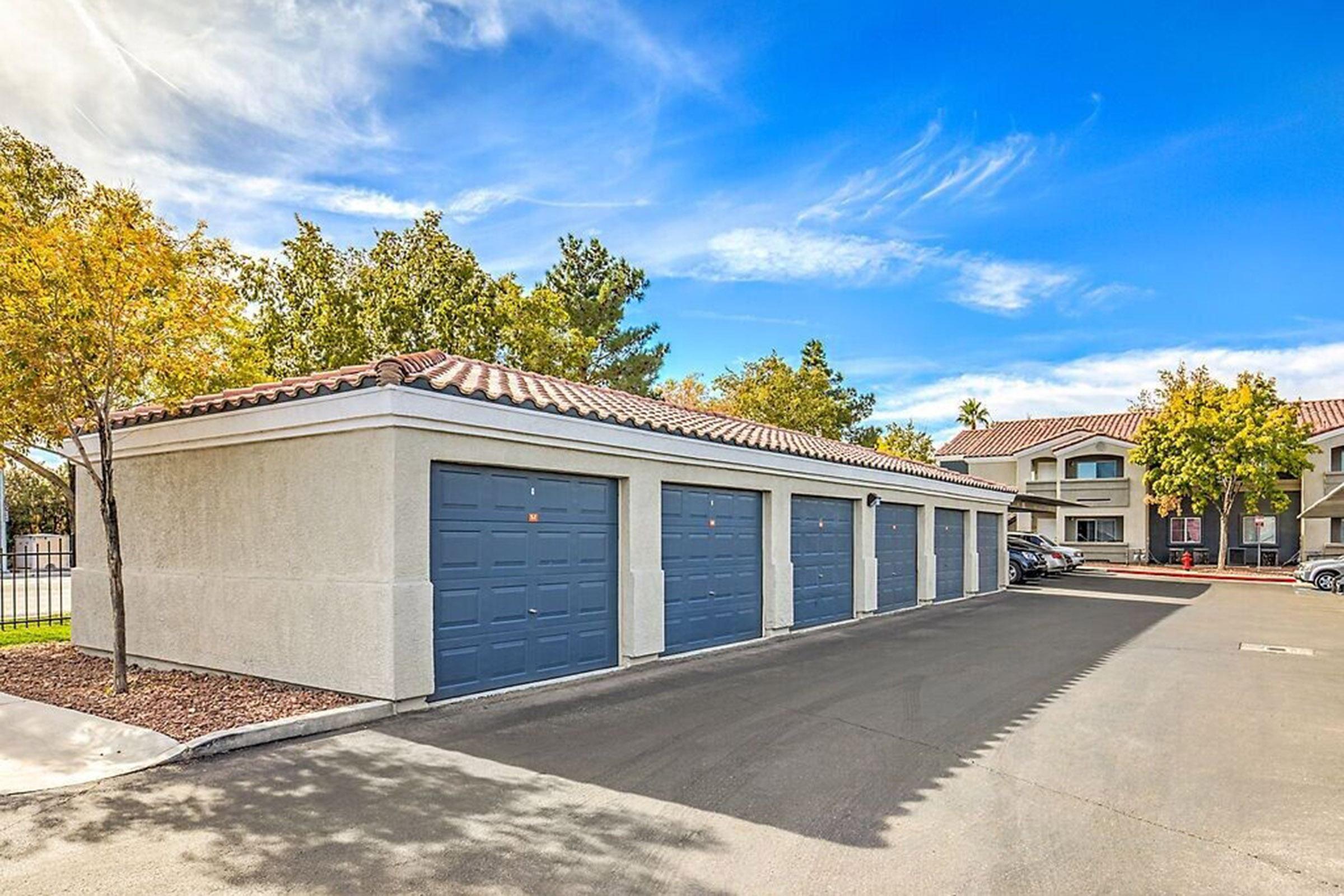 A row of blue garage doors in a well-maintained area, surrounded by trees with fall foliage. The sky is bright blue with wispy clouds. Nearby buildings are visible, indicating a residential complex. The asphalt driveway appears clean and recently paved.