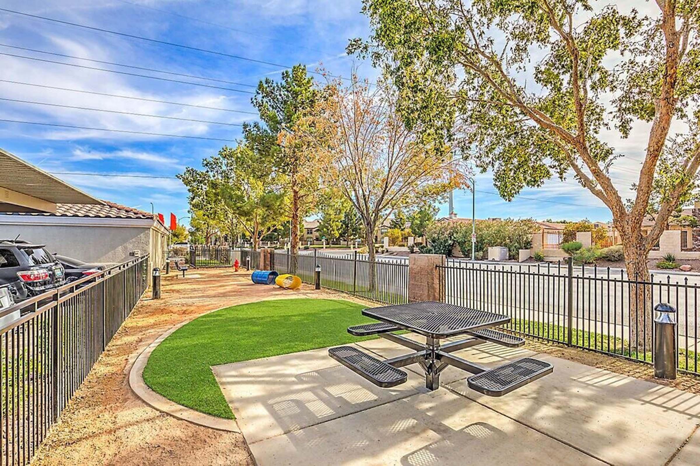 A landscaped outdoor area featuring a grassy space with a picnic table and colorful playground equipment in the background. Trees provide shade, and a fence encloses the area. The sky is clear with a few clouds, creating a pleasant atmosphere.